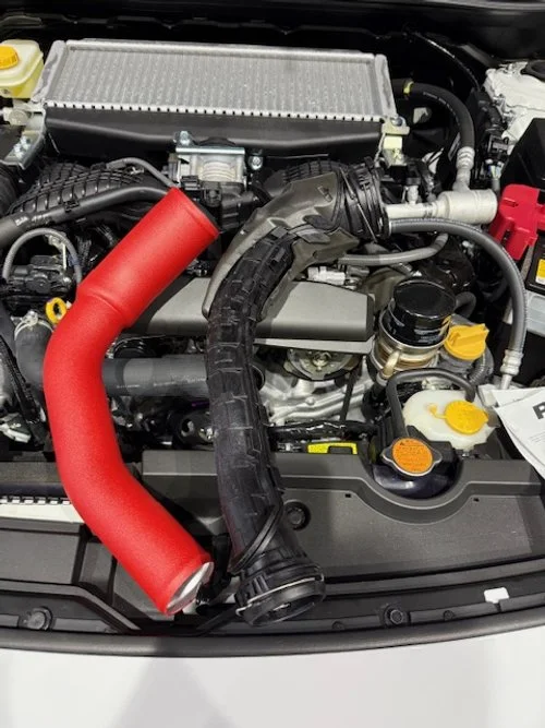 Automobile engine bay with various components including a red intake pipe and black hoses, visible battery, and fluid reservoirs.