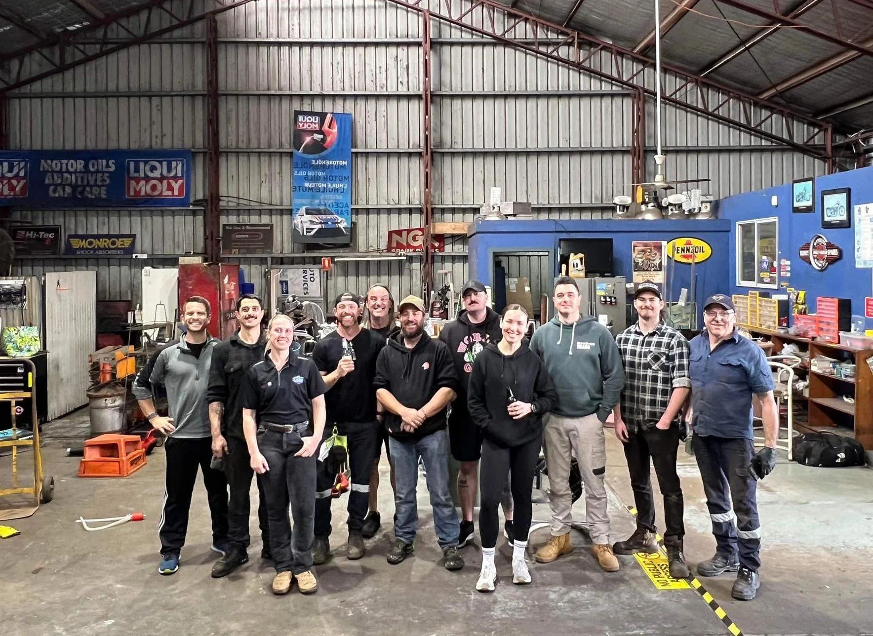 Group of people standing in an auto workshop, with tools and equipment visible in the background.