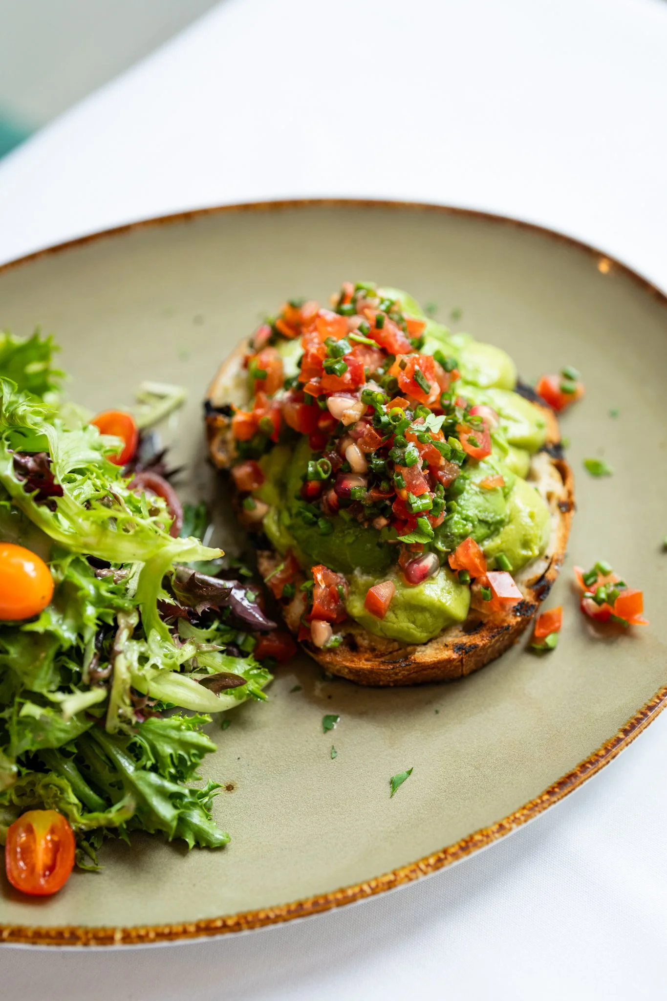 A close-up of a plate with a grilled vegetable dish featuring a halved roasted potato topped with avocado and diced tomatoes, garnished with chives, served alongside mixed green salad with cherry tomatoes.