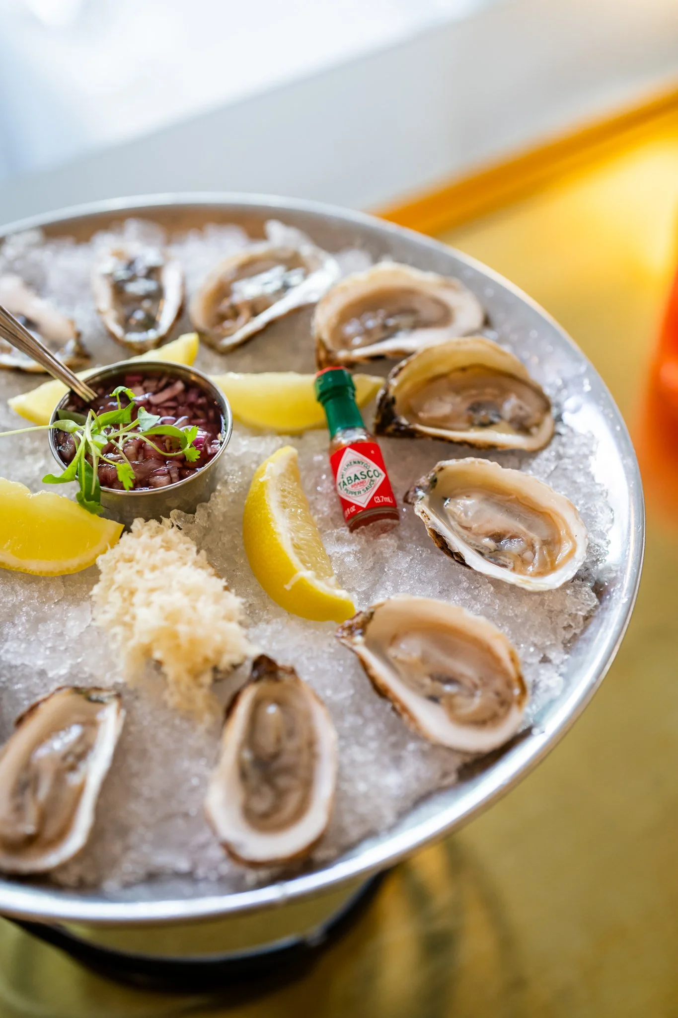 A plate of fresh oysters on ice with lemon wedges, a small bowl of red beans, a sprig of cilantro, grated horseradish, and a small bottle of Tabasco sauce.