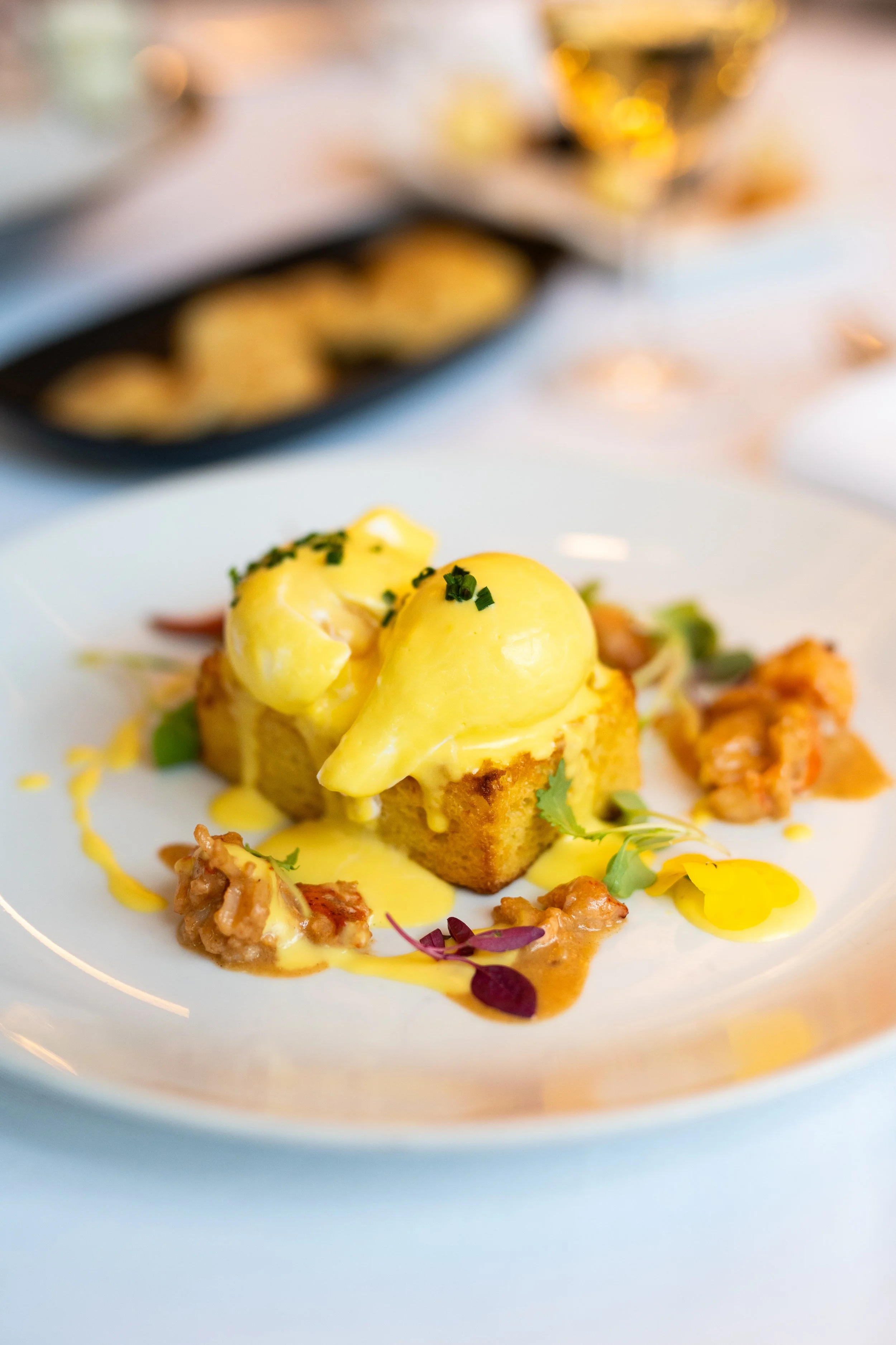 A plated gourmet dish featuring a square piece of cake topped with two scoops of yellow ice cream, garnished with chopped herbs and microgreens, with a drizzle of yellow and brown sauce, and small bits of meat or seafood on a white plate with a blurred background of additional food items.