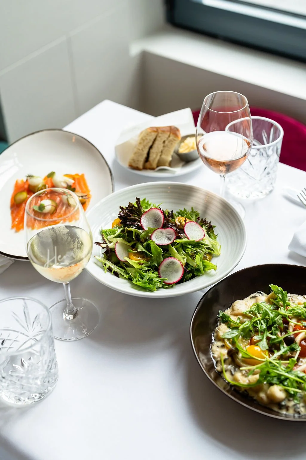 A table with a salad topped with radishes, a glass of white wine, a bowl of pasta with greens, a plate of smoked salmon, a bread basket, and glasses of water.
