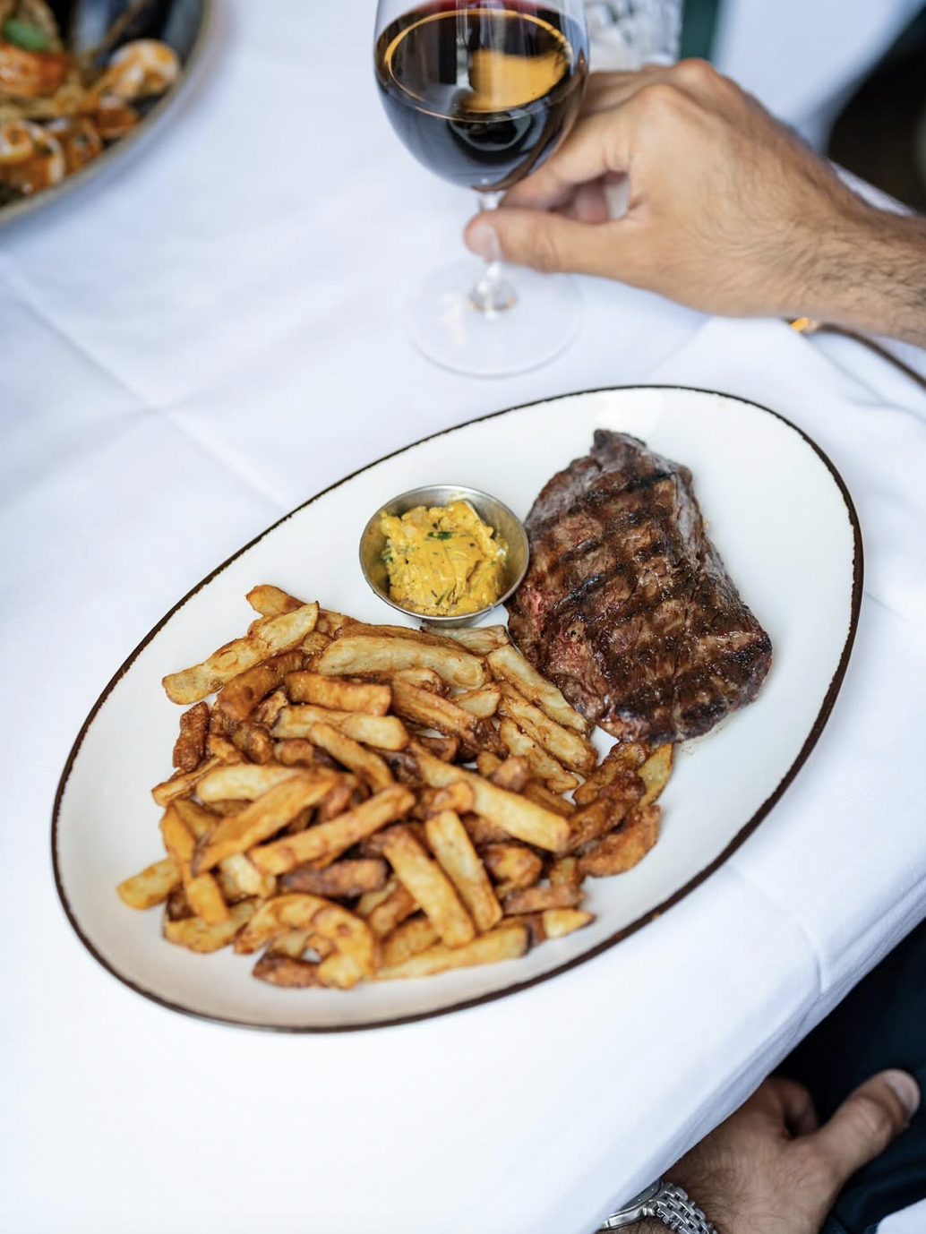 Plate with French fries, a grilled steak, and a small dish of yellow mustard or dipping sauce, with a glass of red wine above the plate.