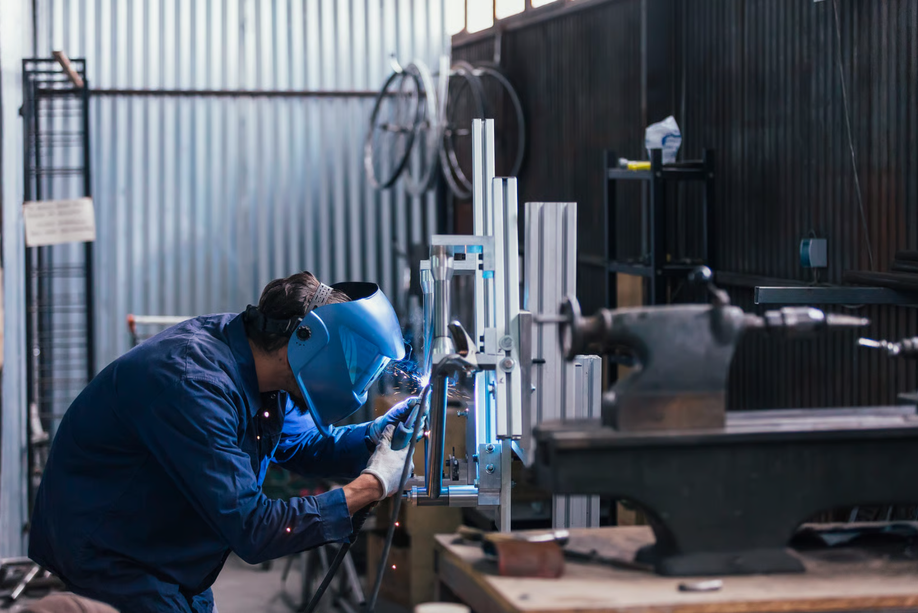 A person wearing a welding helmet and blue work clothes welding metal components in a workshop.