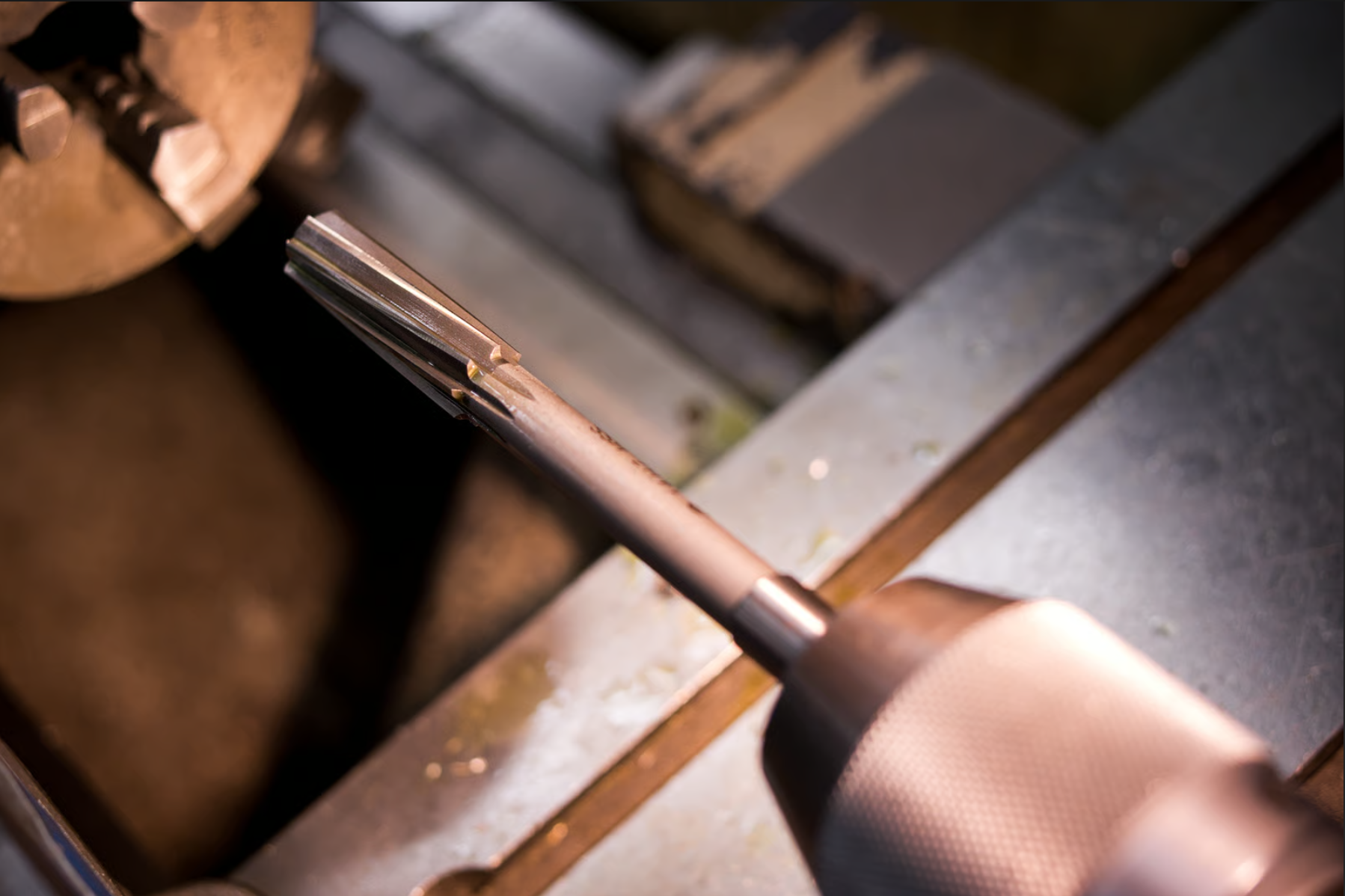 Close-up of a metal tool with a flat, beveled edge being used on a workbench. The workspace has metal and wooden parts, indicating a workshop or fabrication setting.