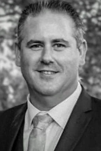 Black and white portrait of a man in a suit and tie, smiling, outdoors on a cloudy day.