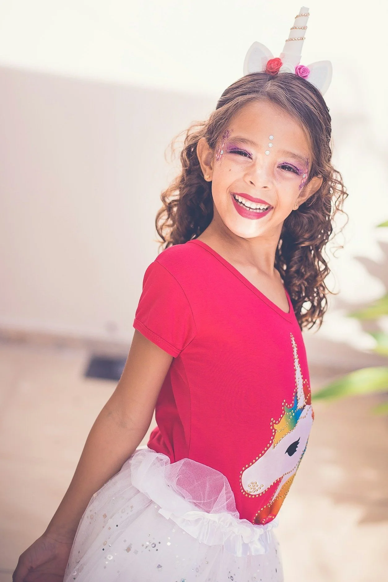 A young girl dressed as a unicorn princess, smiling, with unicorn horn headband, colorful makeup, and a pink shirt with a unicorn graphic, wearing a white tutu