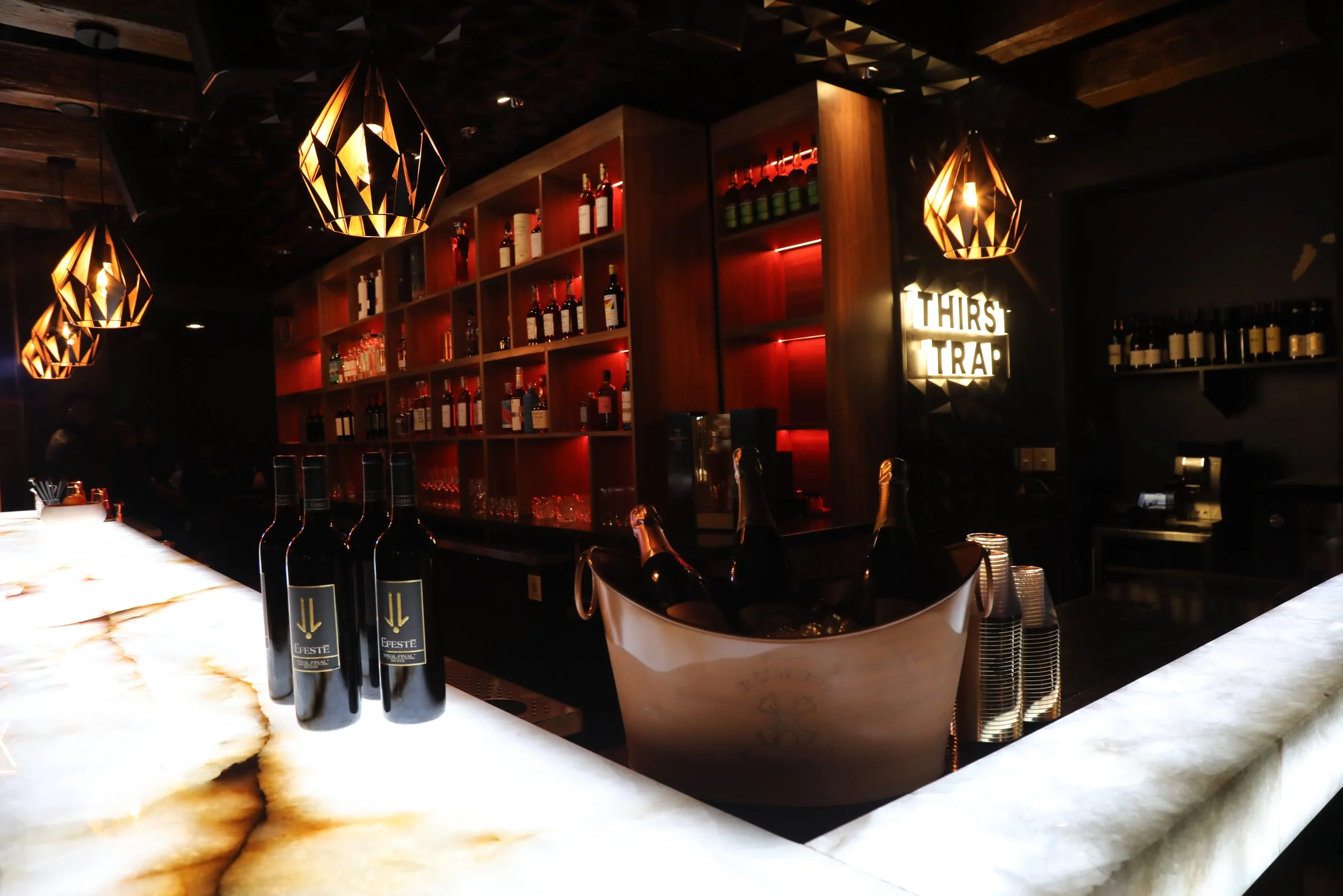 Dimly lit bar interior with bottles on shelves, hanging light fixtures, "Thirst Trap" sign, and wine bottles on a marble countertop.