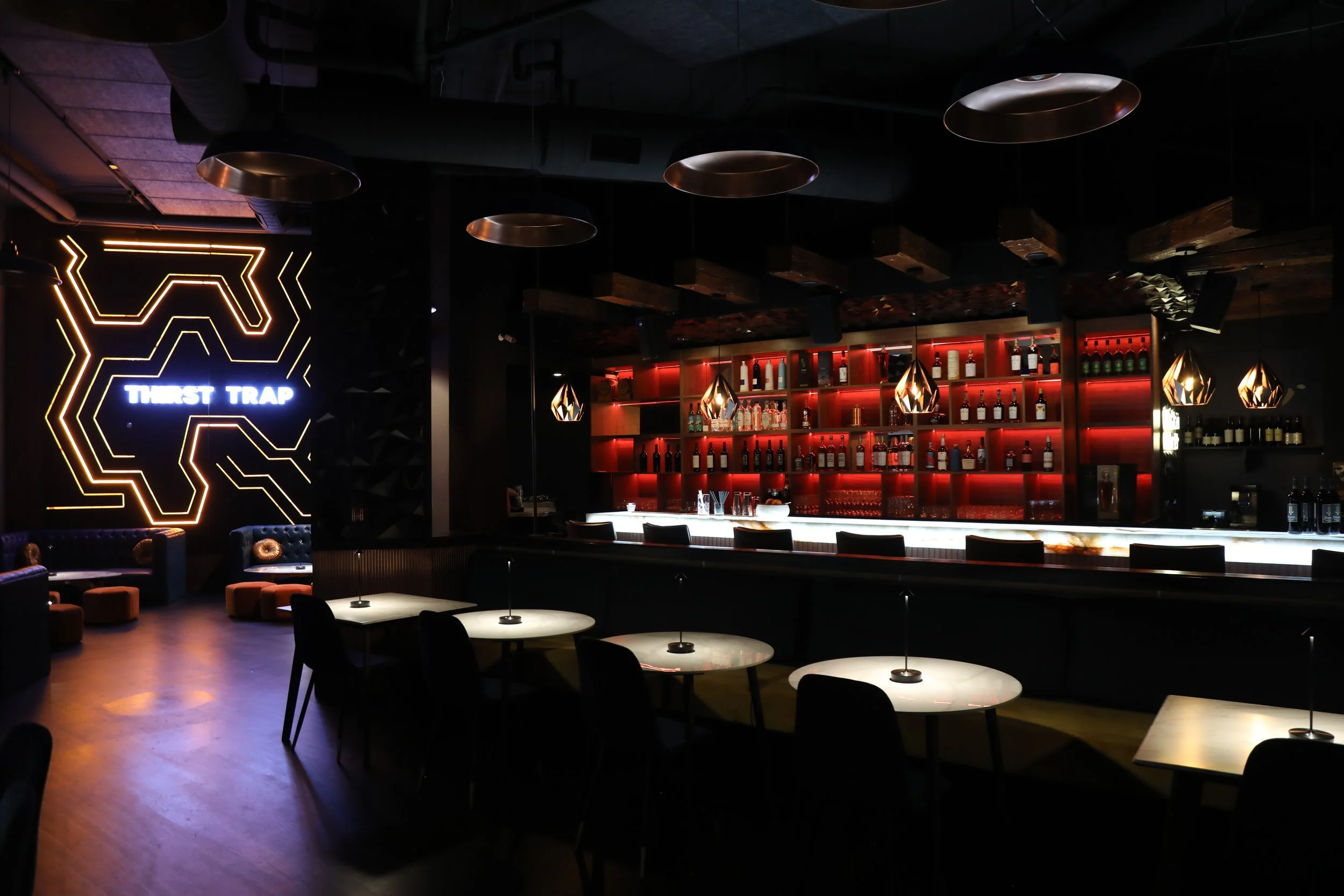 Modern bar interior with illuminated shelving, bottles of liquor, pendant lights, tables, and a sign reading 'Thirst Trap.'