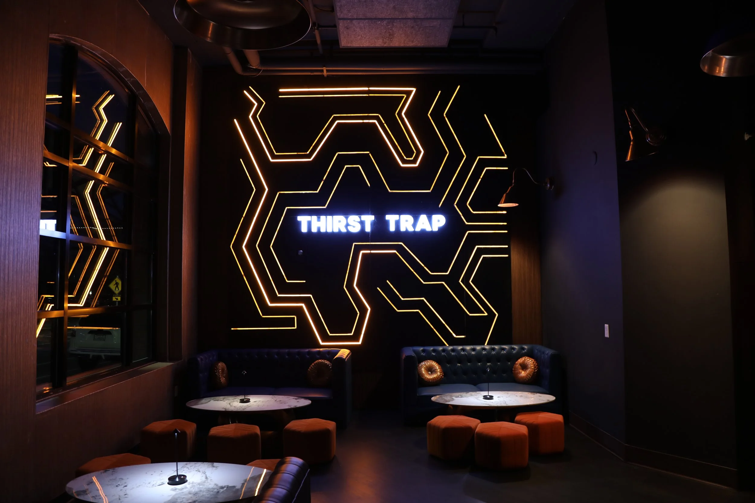 Modern lounge interior with neon lights and 'Thirst Trap' sign, featuring round tables, orange stools, and black cushioned seating.