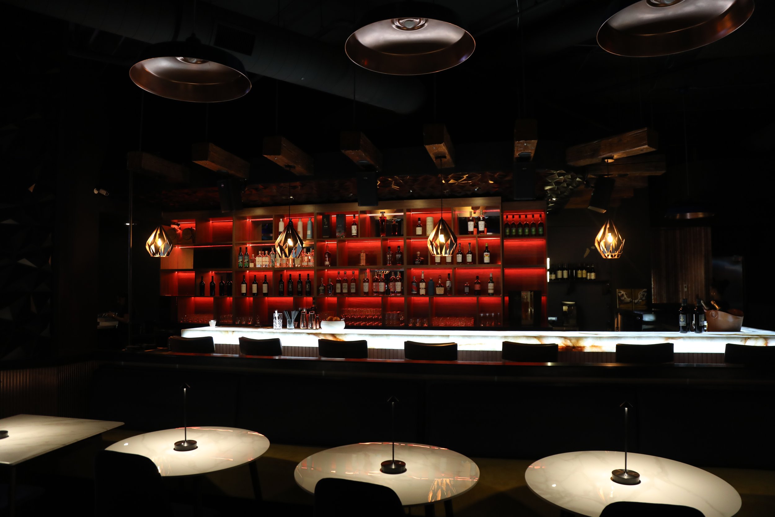 Dimly lit modern bar with red backlit shelves and hanging lights.