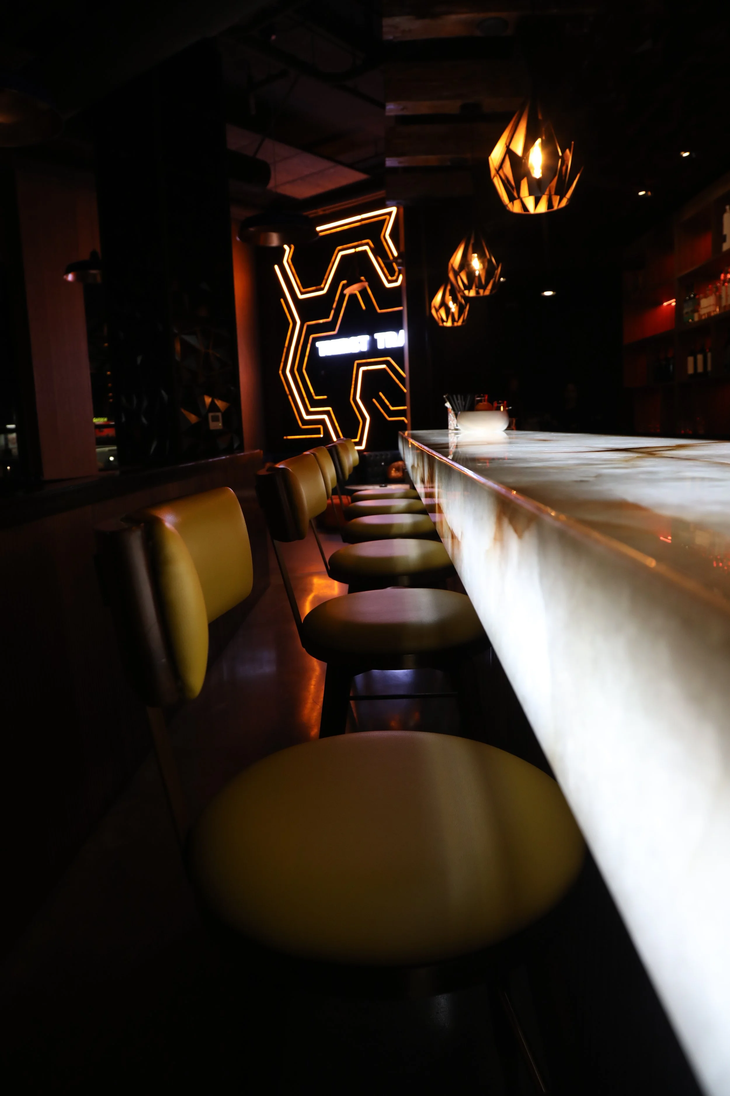 Modern dimly lit bar interior with yellow cushioned stools, sleek white bar counter, decorative geometric lighting, and neon wall art.