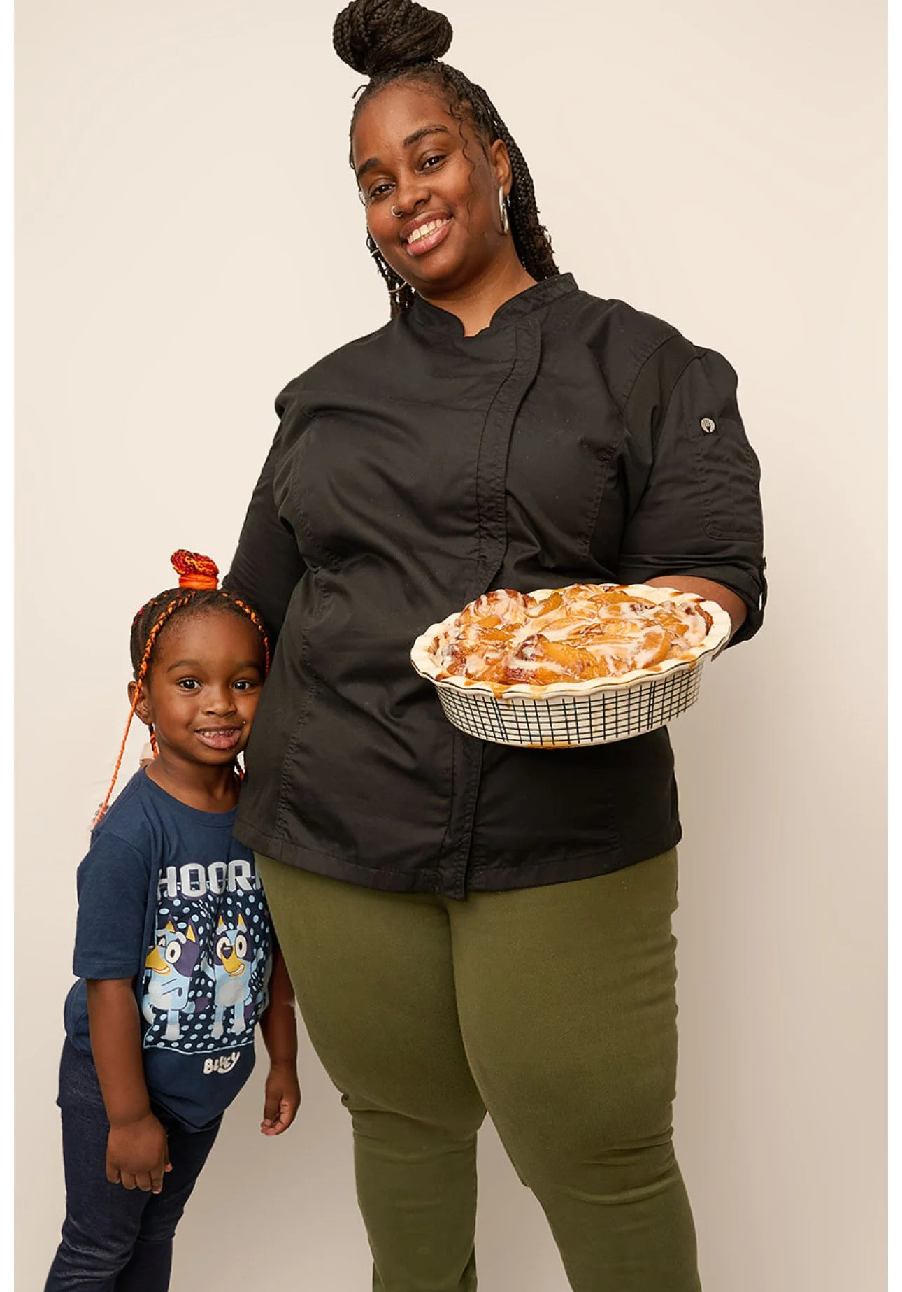 A woman with dark hair in braids and a bun, wearing a black chef's coat, holding a pie, smiling. A young girl with braided hair and colorful hair accessories, wearing a blue shirt with cartoon characters, standing beside her, smiling.
