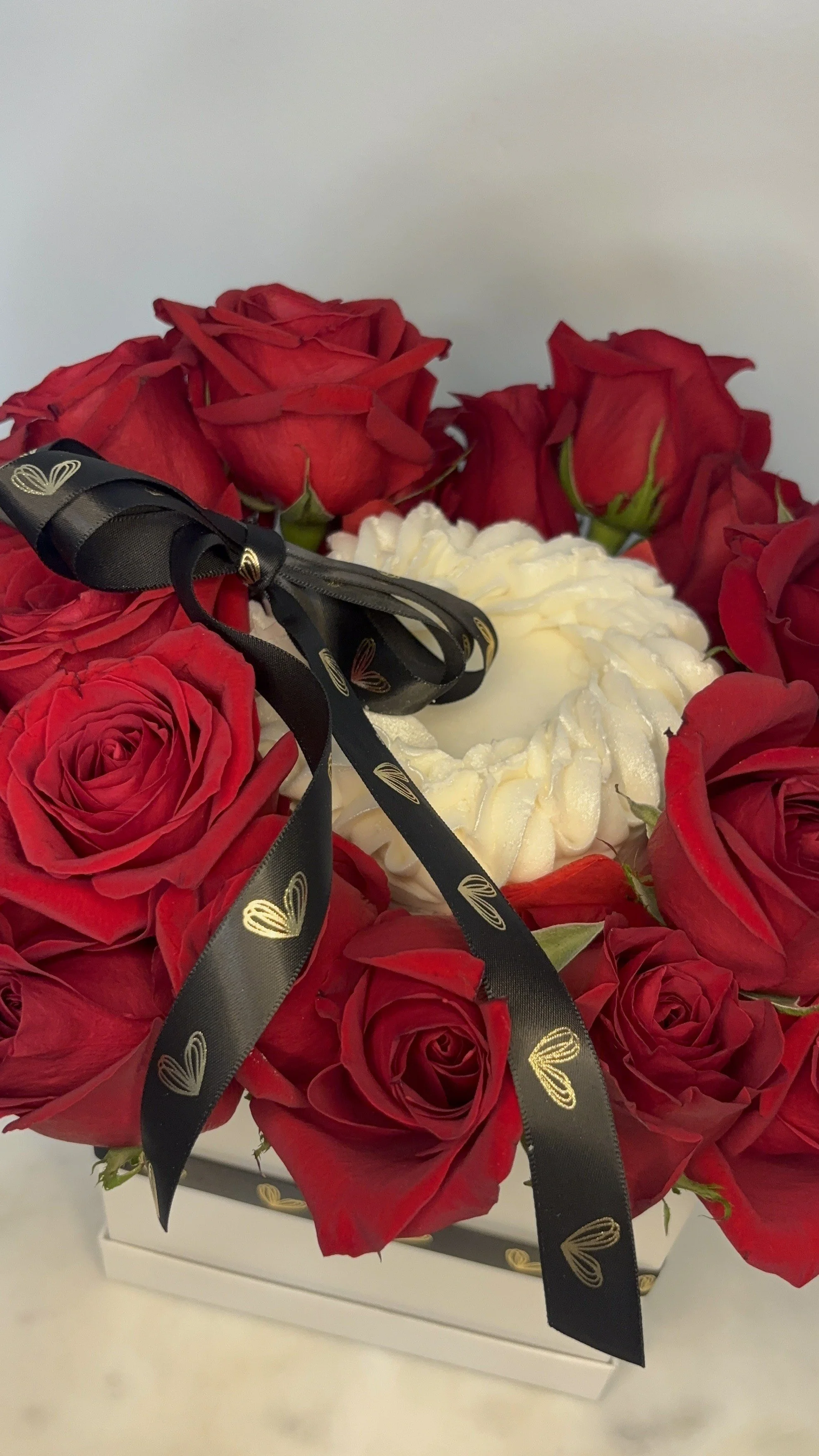 A bouquet of red roses surrounding a heart-shaped cake with white frosting, adorned with a black ribbon with gold heart patterns, in a white box.