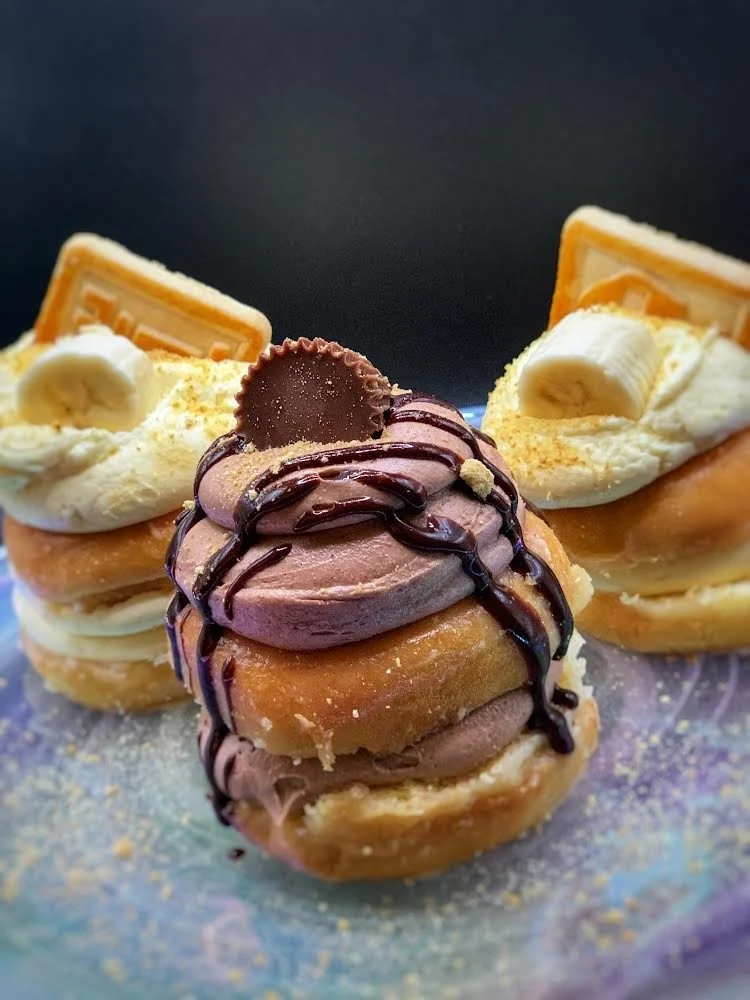 Close-up of three cream-filled donuts topped with chocolate, whipped cream, and banana slices, garnished with chocolate drizzles and biscuit pieces.