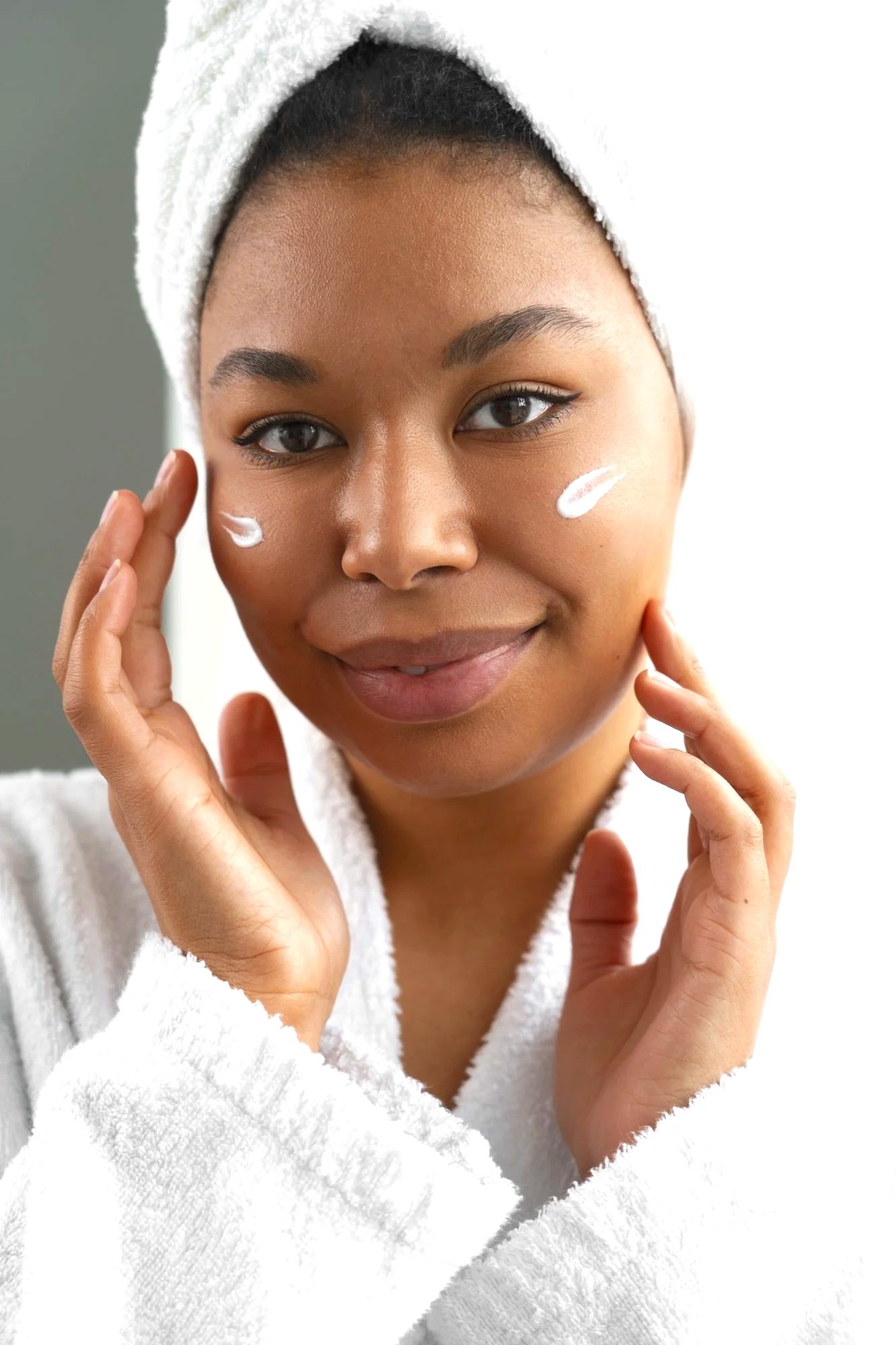 A woman in a white towel wrapping her head, applying skincare cream to her cheeks, smiling confidently.