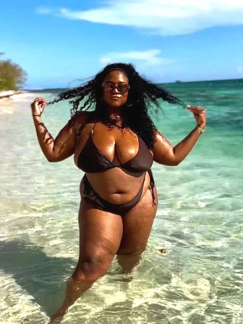 A woman in a dark brown bikini with cutouts standing knee-deep in clear ocean water at the beach, holding her curly hair, with a blue sky and distant shoreline in the background.