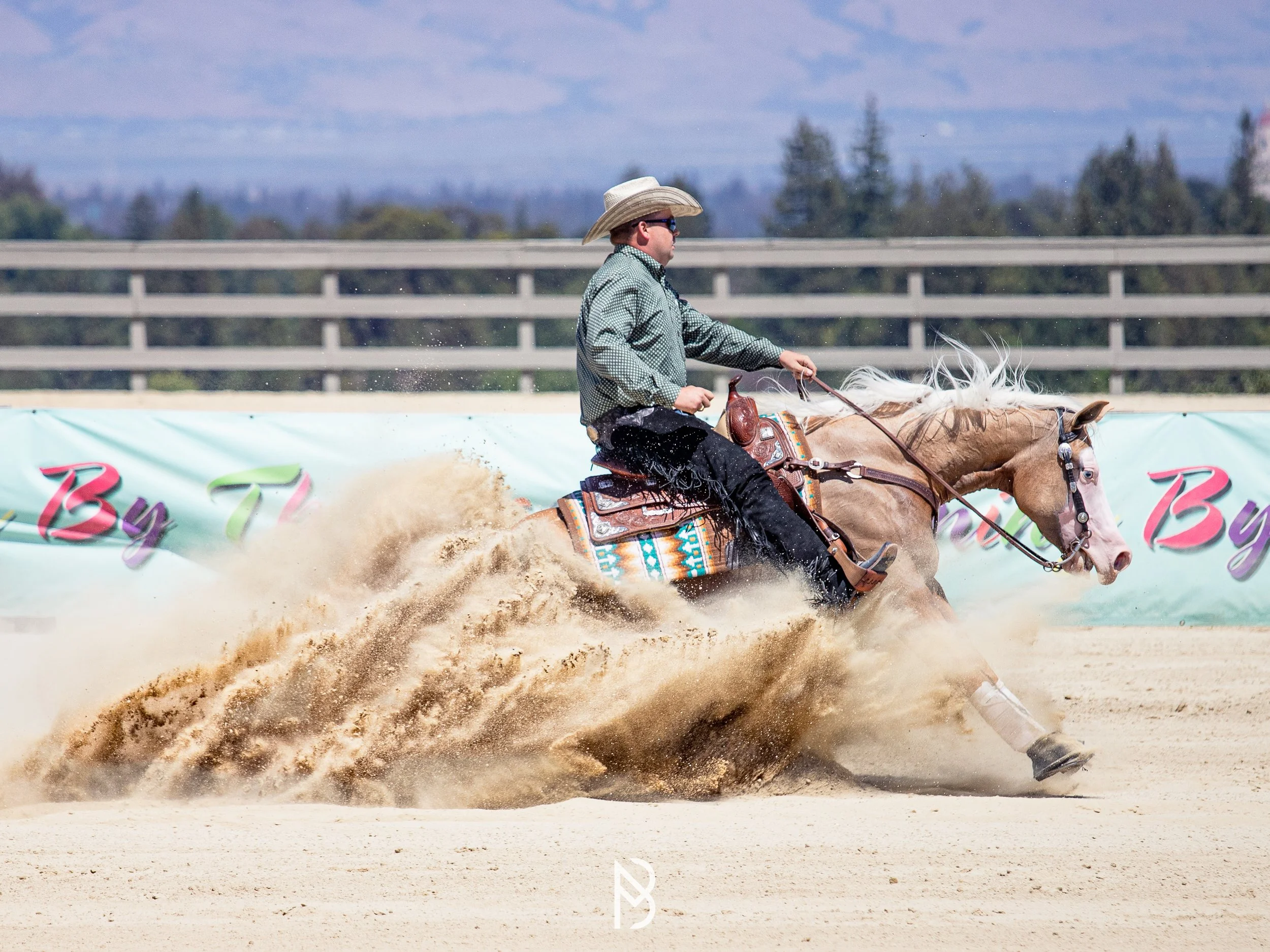 A cowboy riding a light brown horse at a gallop, kicking up dust on a dirt arena.