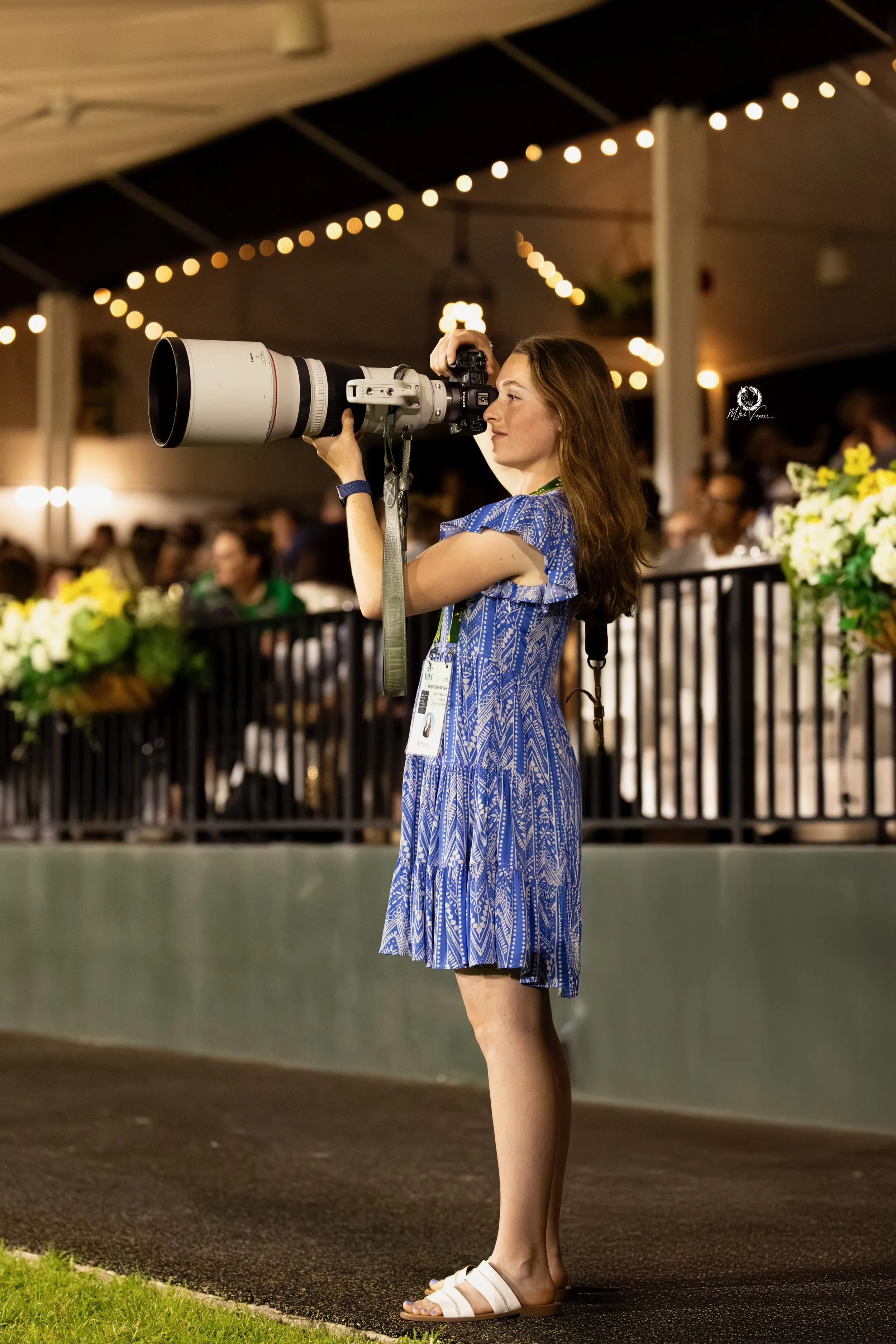 A woman in a blue patterned dress takes photographs with a large camera at an indoor event decorated with string lights and floral arrangements.
