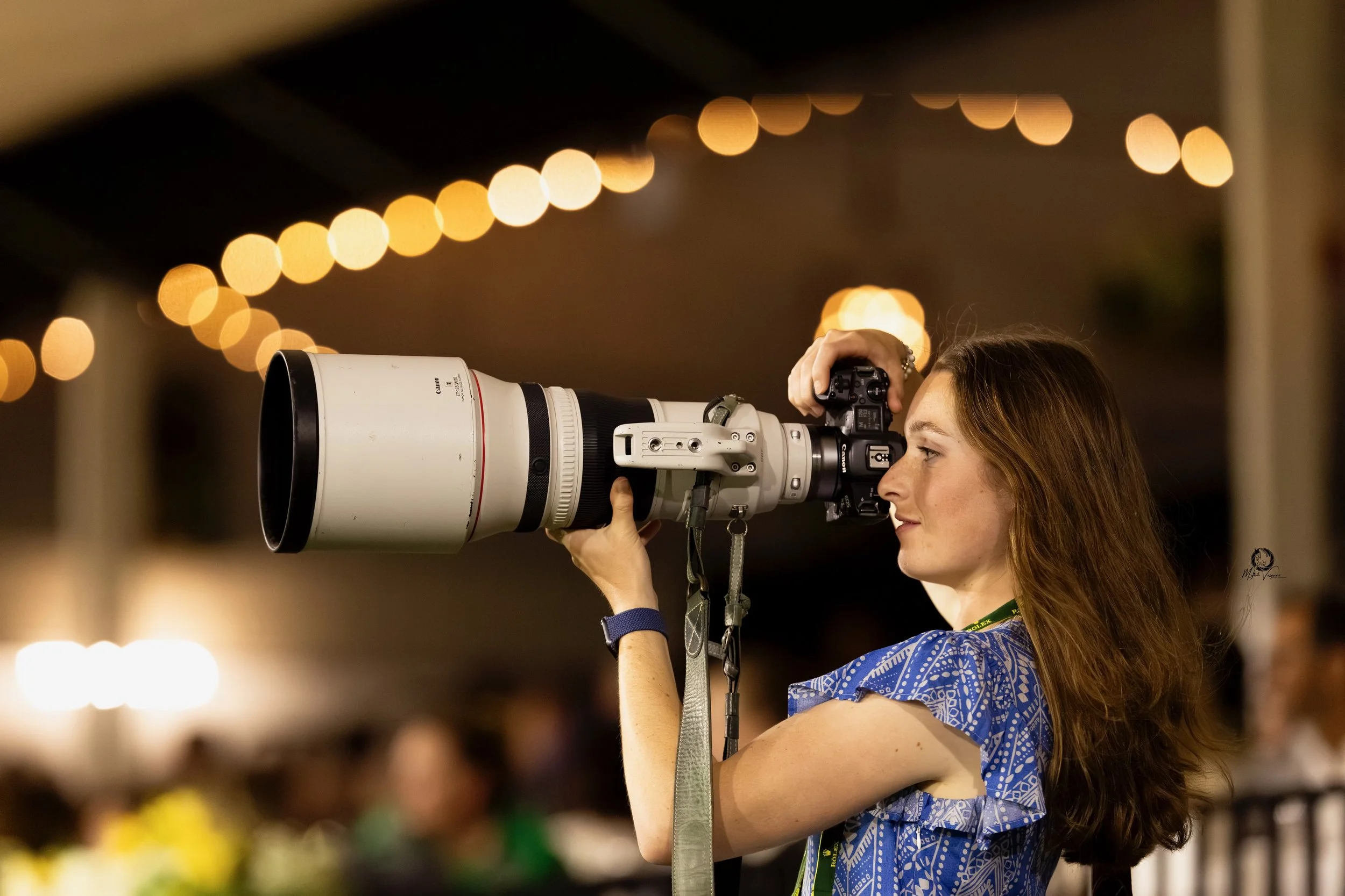 A woman with long hair in a blue patterned dress takes a photograph with a large telephoto camera lens during an indoor event with warm string lights in the background.