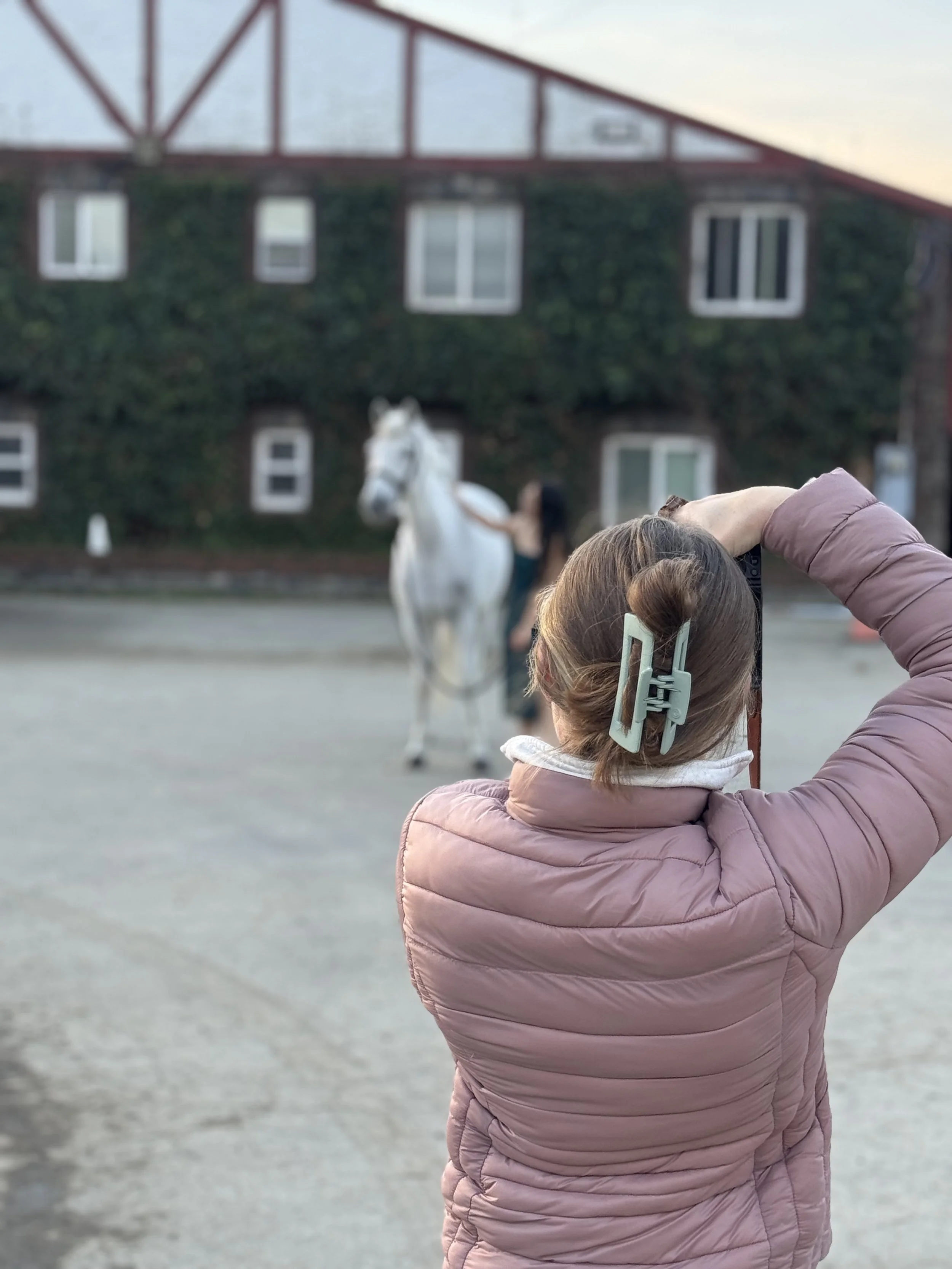 A person with brown hair in a bun, wearing a pink jacket, is adjusting their hair with a clip while facing a white horse. The horse is being held by a woman in the background, with a green, ivy-covered building behind them.
