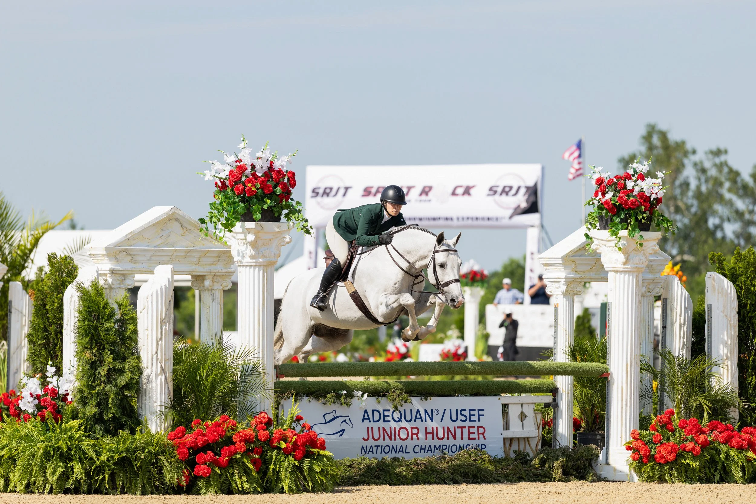A rider on a white horse jumping over an obstacle during a show jumping competition at the Adequan USEF Junior Hunter National Championship, in Lexington Kentucky, decorated with red and white flowers and white columns.
