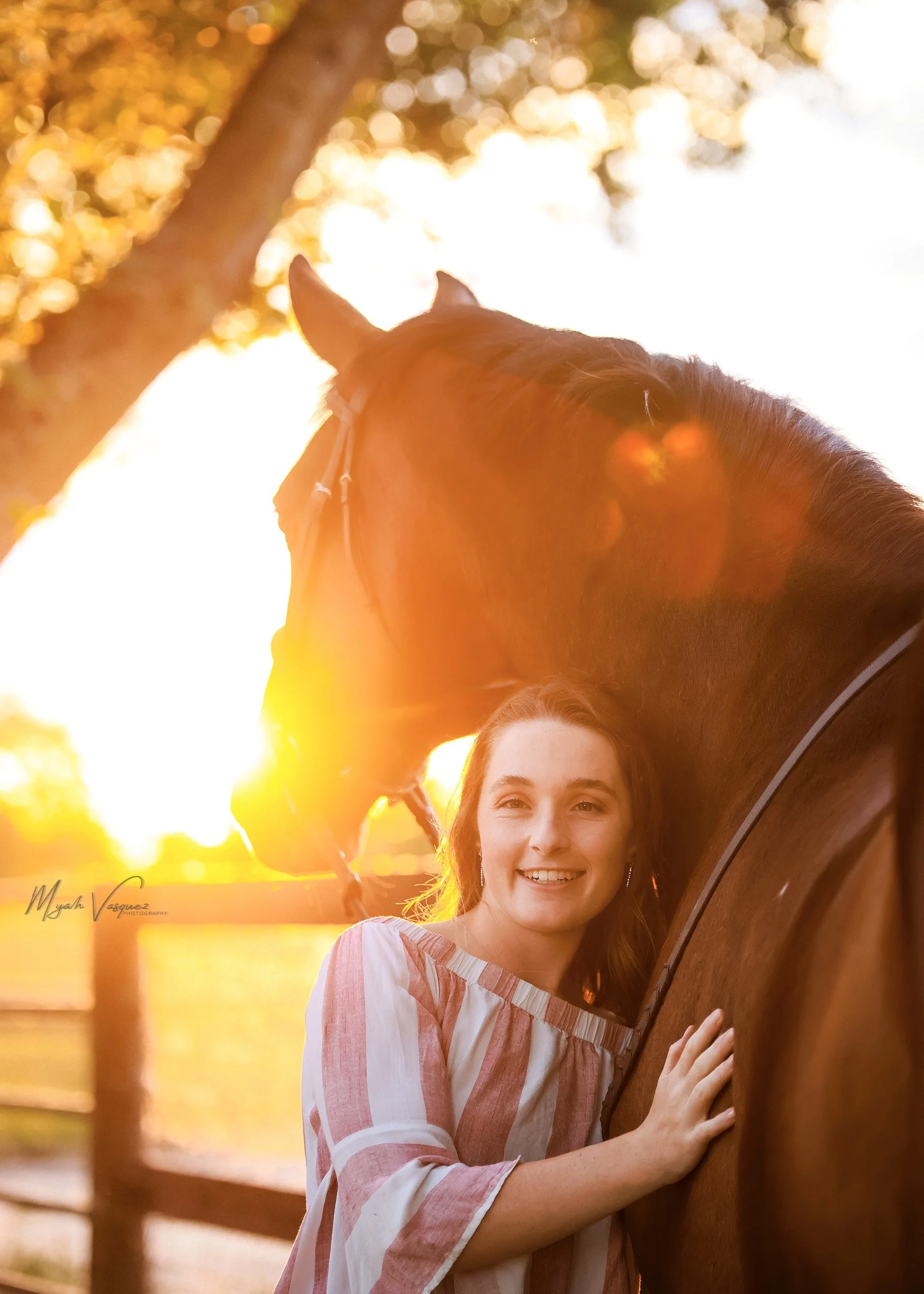 A woman smiling and petting a horse during sunset, with sunlight shining through trees in the background.