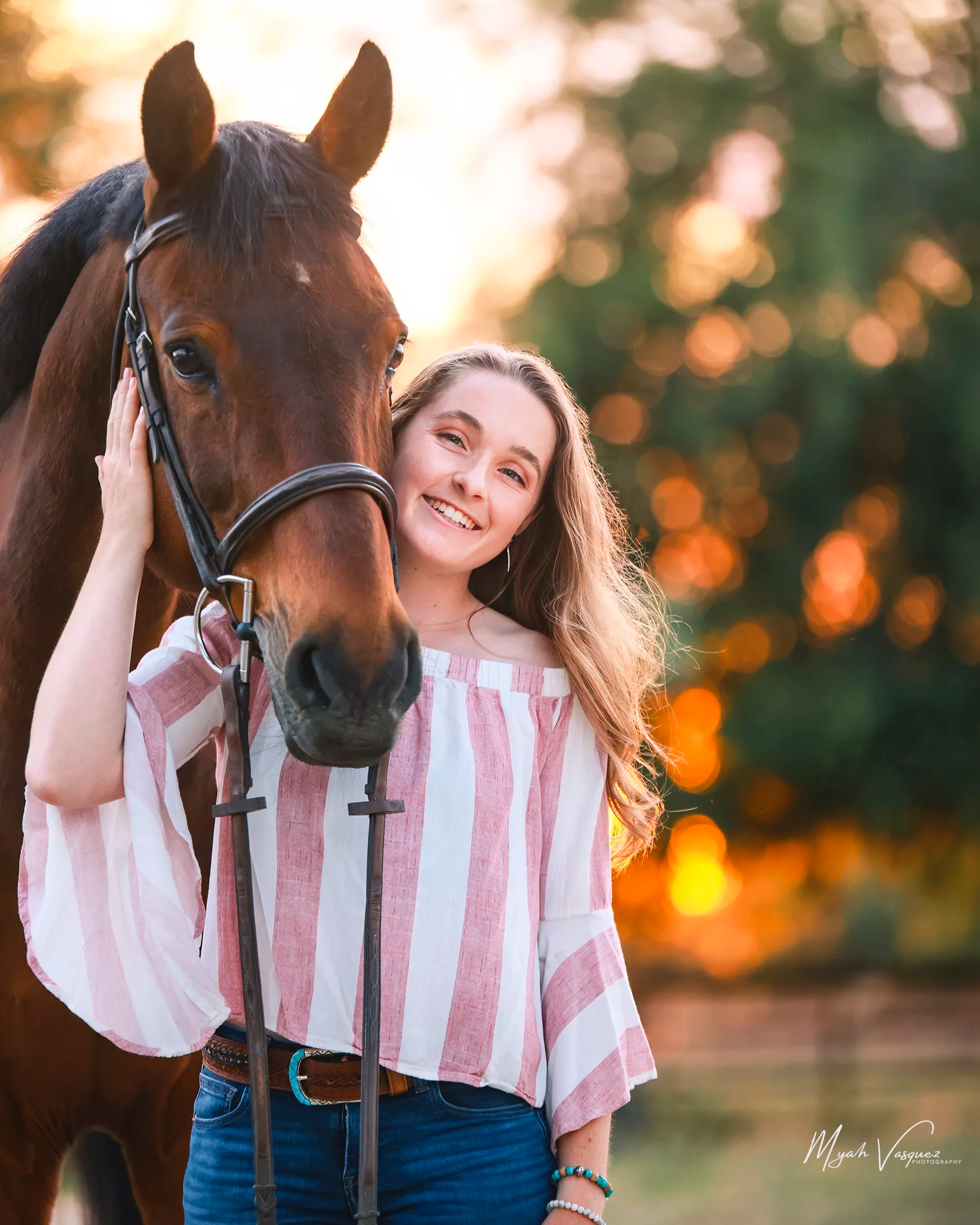 A young woman with long blonde hair smiling and hugging a brown horse outdoors during sunset, with a background of orange and green trees.