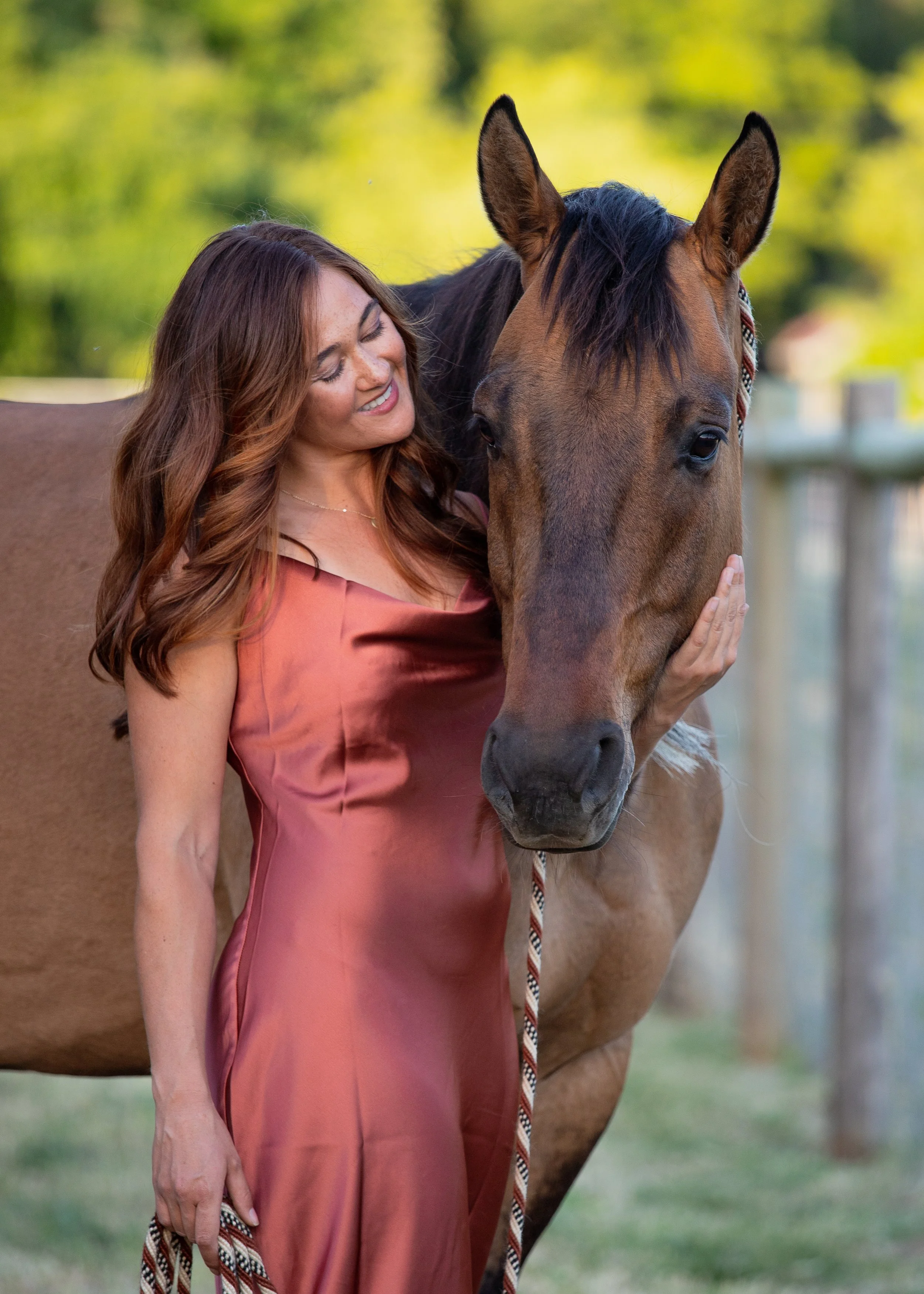 A woman with long, wavy auburn hair wearing a silky, rust-colored dress, smiling and gently touching a brown horse's face outdoors in a grassy area with trees in the background.