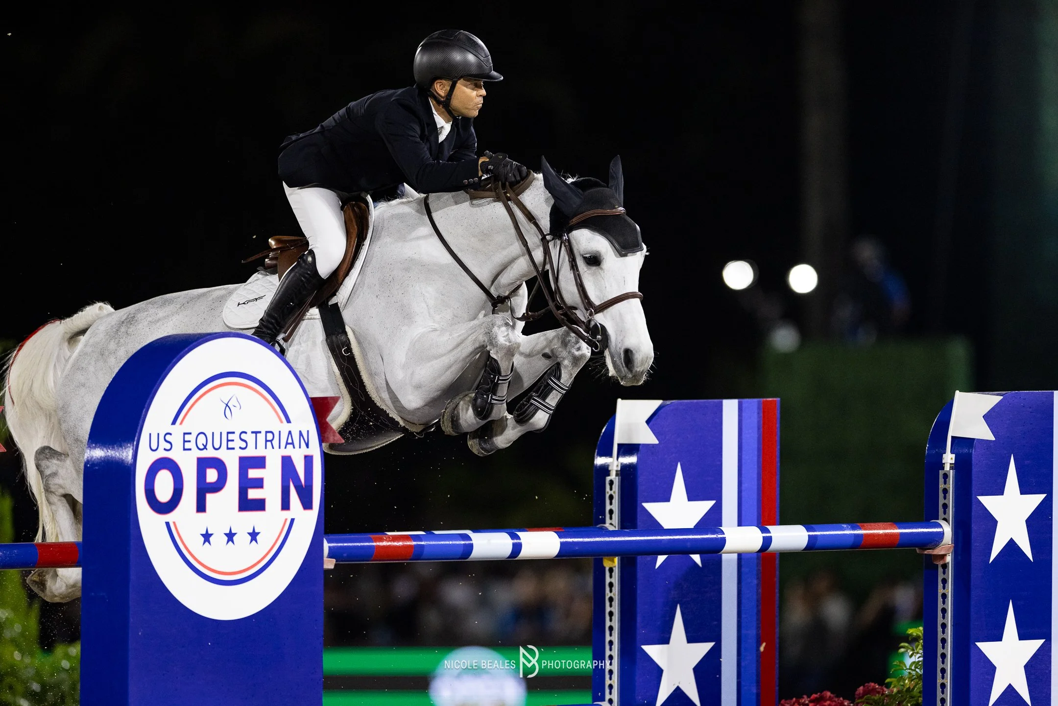 A Kent Farrington and Greya jumping over an obstacle at a nighttime equestrian event in Wellington Floriday, with a sign reading 'US Equestrian Open' on the obstacle.