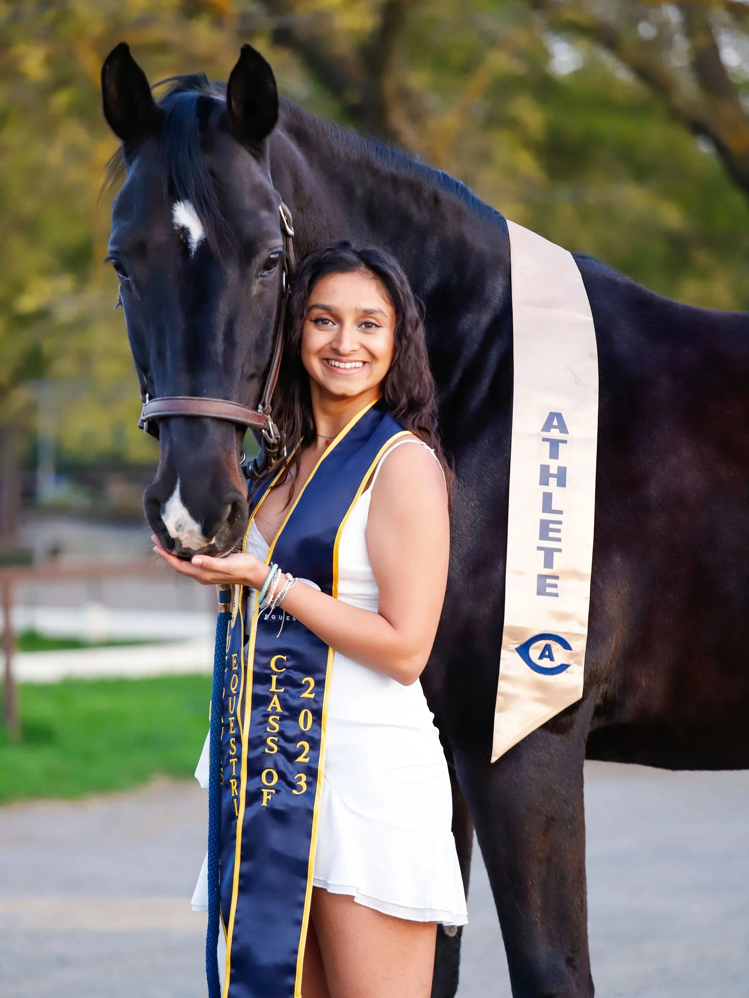 A young woman in a white dress smiles and holds a horse's chin, standing next to a tall black horse with an 'Athlete' sash, outdoors with trees in the background.