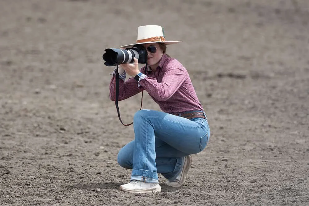 Woman with a camera crouching on a dirt surface, wearing a wide-brimmed hat, sunglasses, a red checkered shirt, blue jeans, and white sneakers.