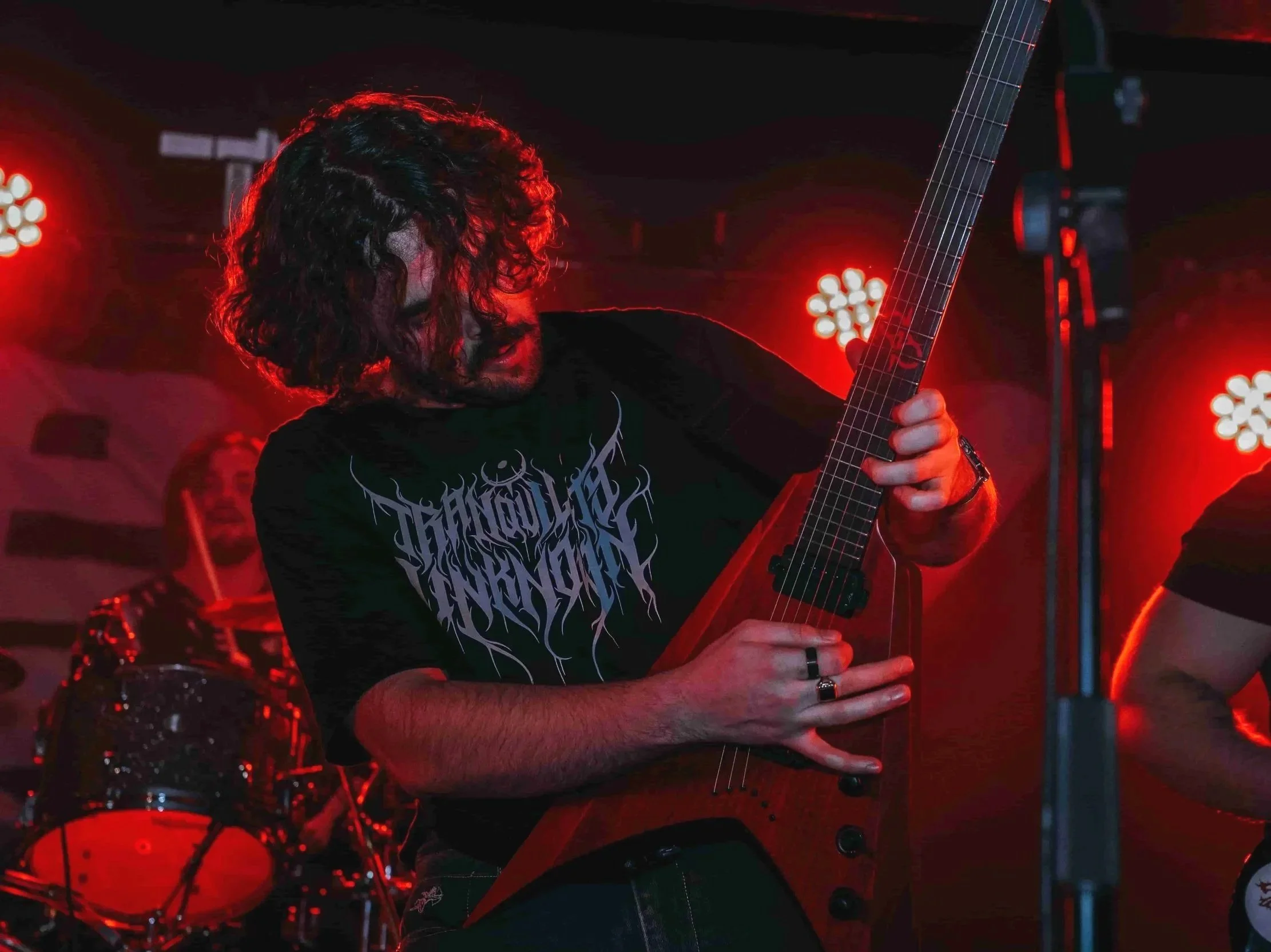 A musician with curly hair is playing an orange electric guitar on stage, illuminated by red stage lights, with a drummer in the background.
