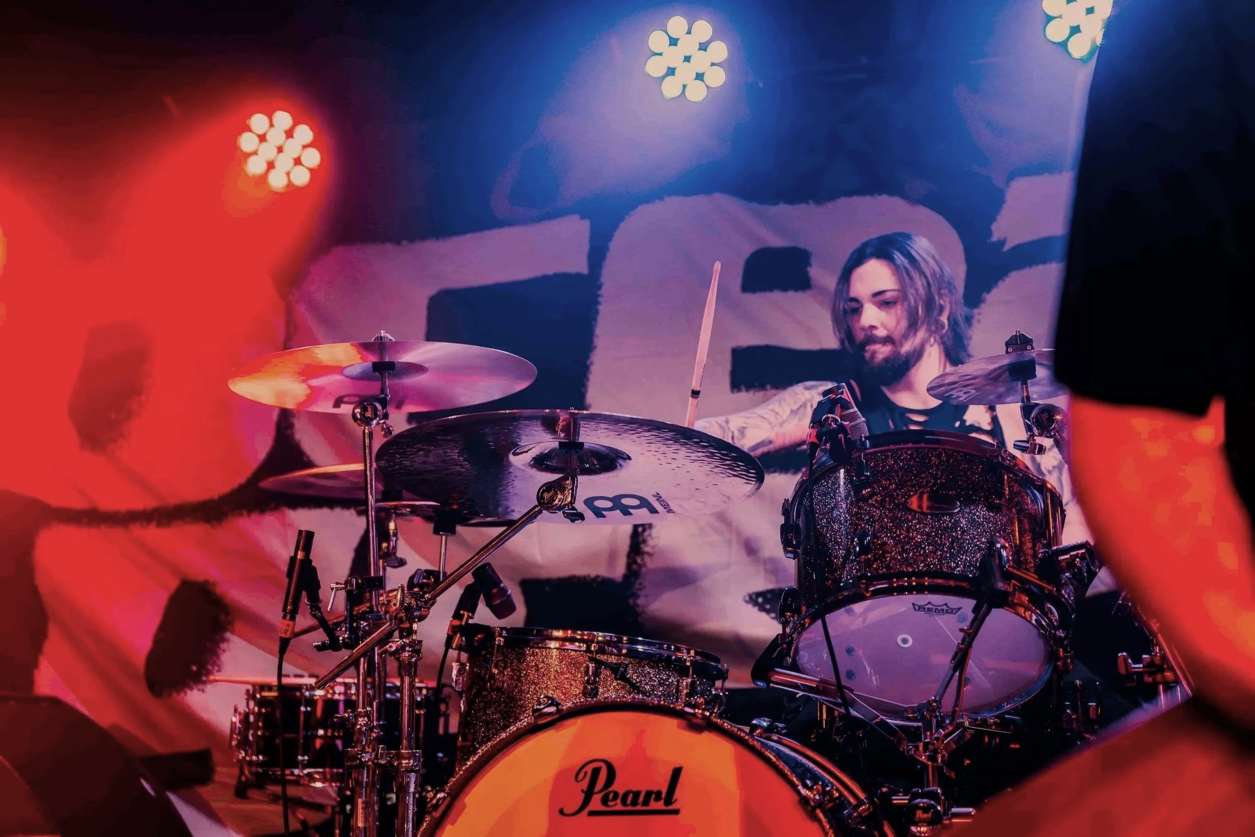 A drummer with long hair and a beard plays a Pearl drum set on stage. The background features a colorful, abstract backdrop with pink, purple, and blue lighting.