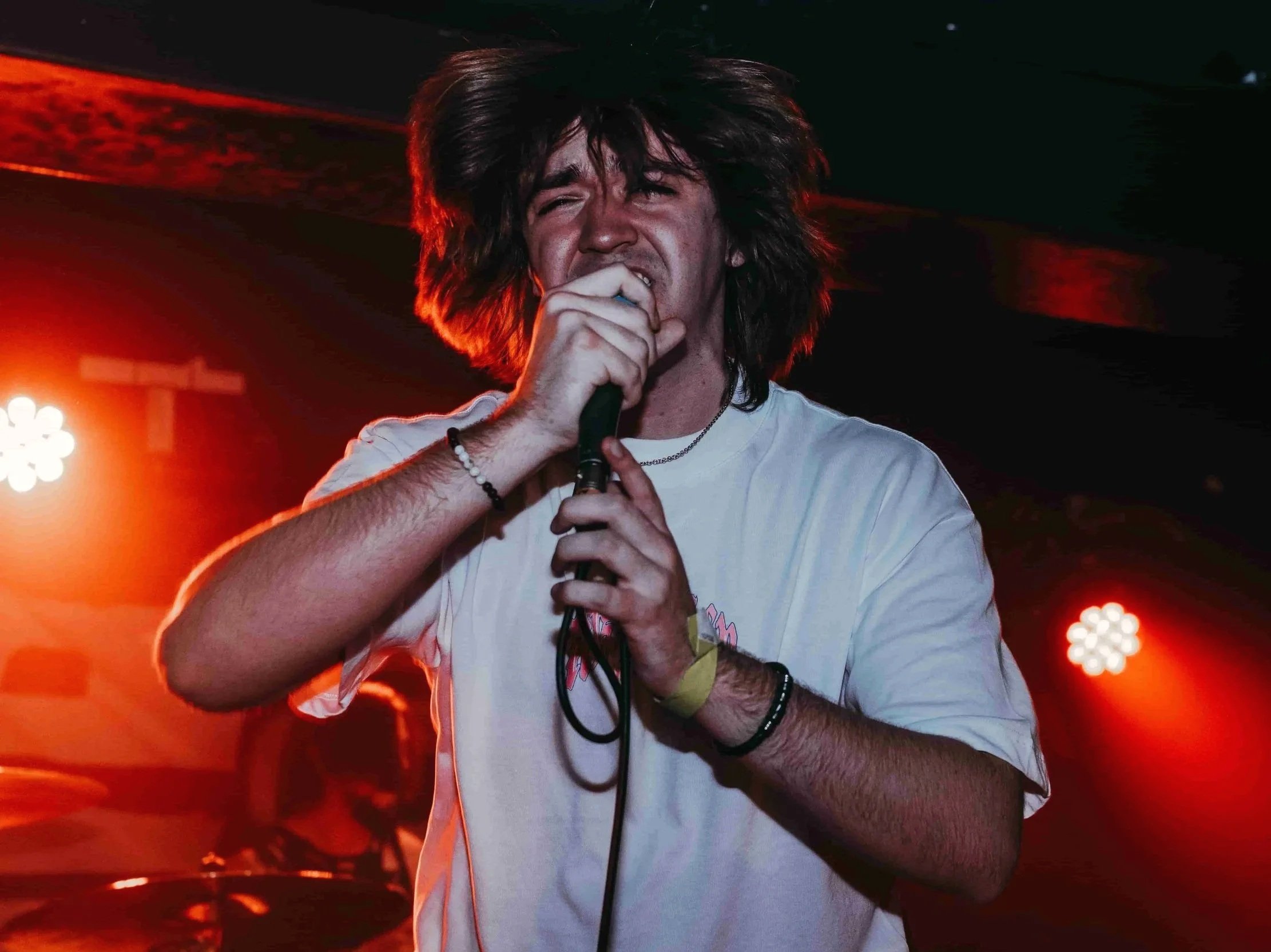 A young man with long, messy hair singing into a microphone on a stage with red and orange lighting.