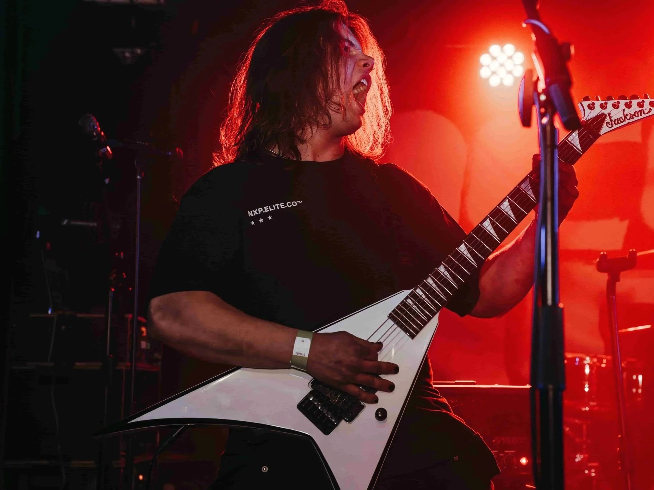 A person with long hair playing a white electric guitar on stage, singing passionately, with red stage lighting and microphones in the background.
