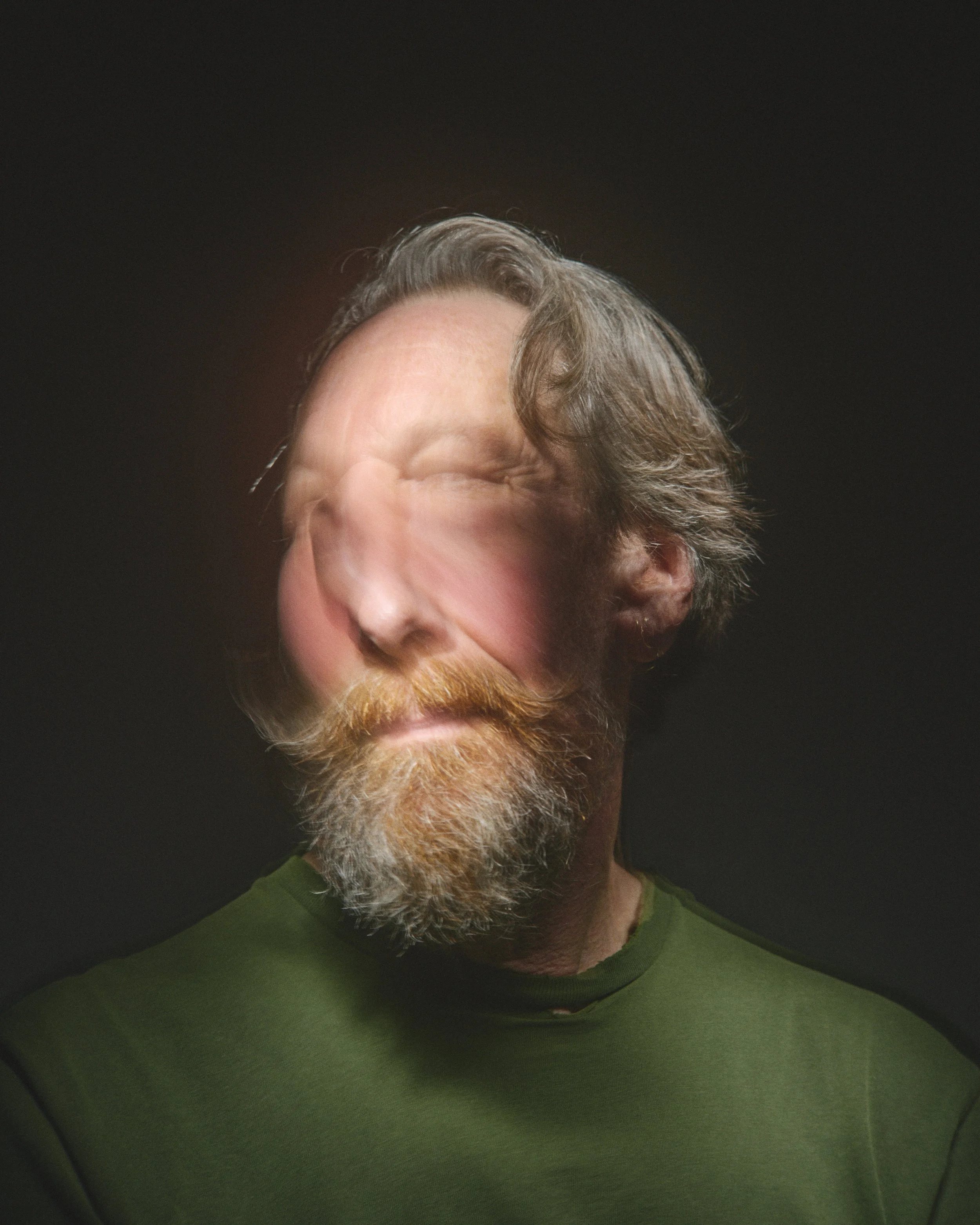 A man's face is distorted with a blurred effect, showing curly hair, a beard, and wearing a green shirt, against a dark background.