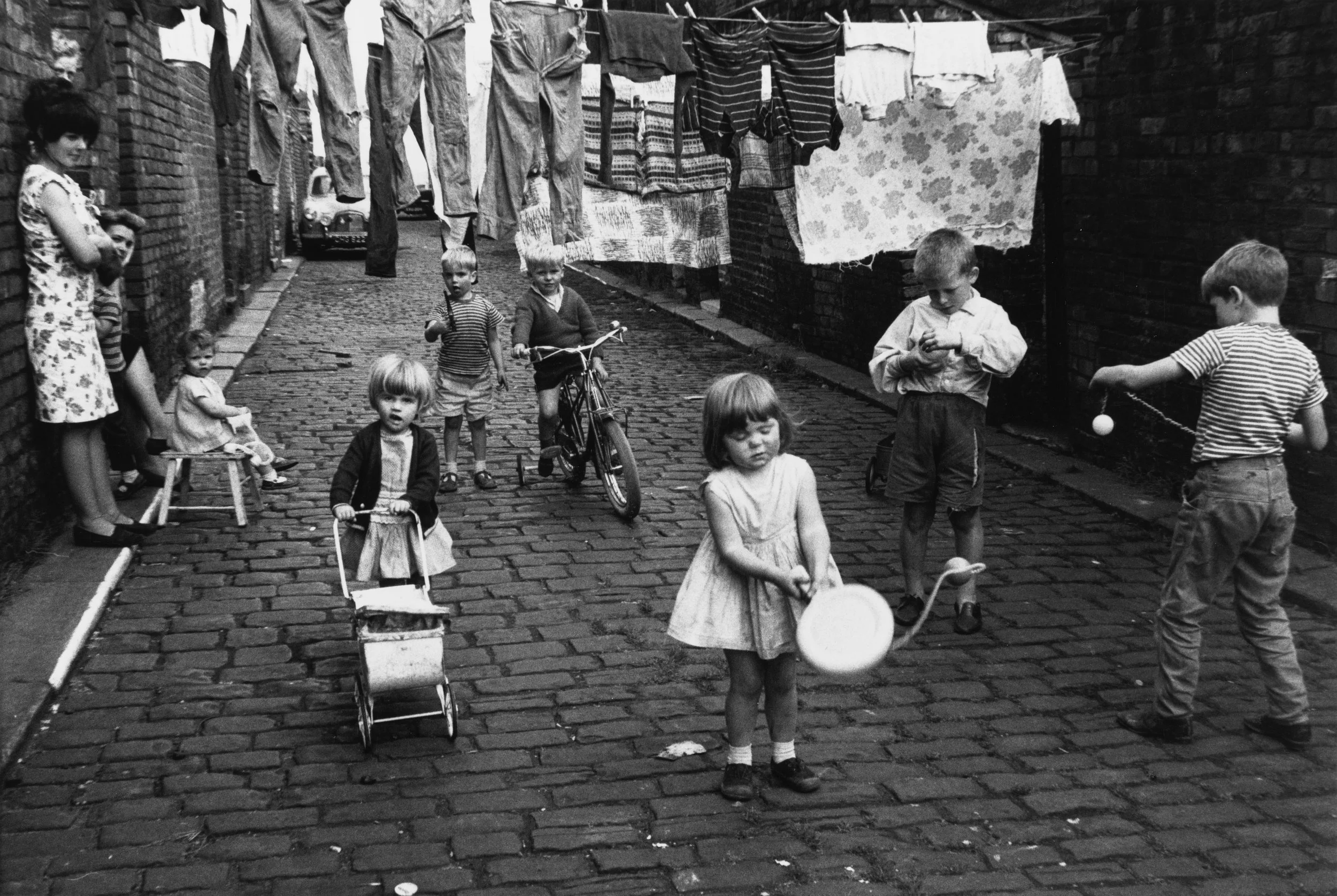 Children playing and socializing in an alleyway with hanging laundry and brick walls, in a black and white photo.