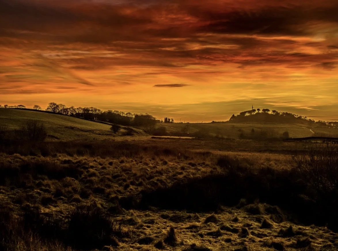 A scenic landscape view at sunset with rolling hills, grassy fields, and a dramatic orange and yellow sky with streaks of clouds.
