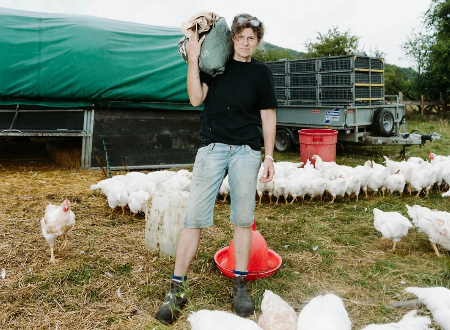 A woman on a farm holding a bag of feed on her shoulder, surrounded by white chickens, with a trailer and trees in the background.