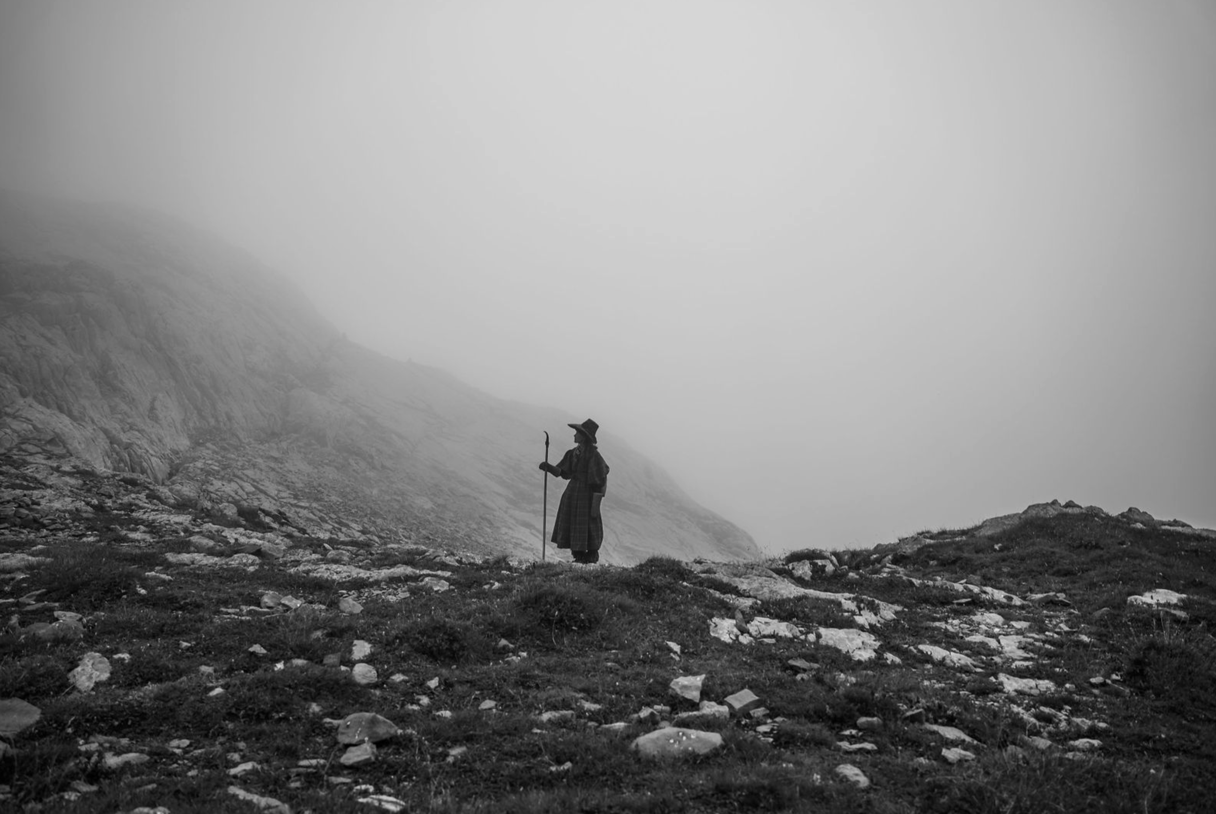 A person dressed in traditional Scottish attire, including a kilt, standing on rocky terrain with a walking stick, surrounded by a foggy mountain landscape in black and white.