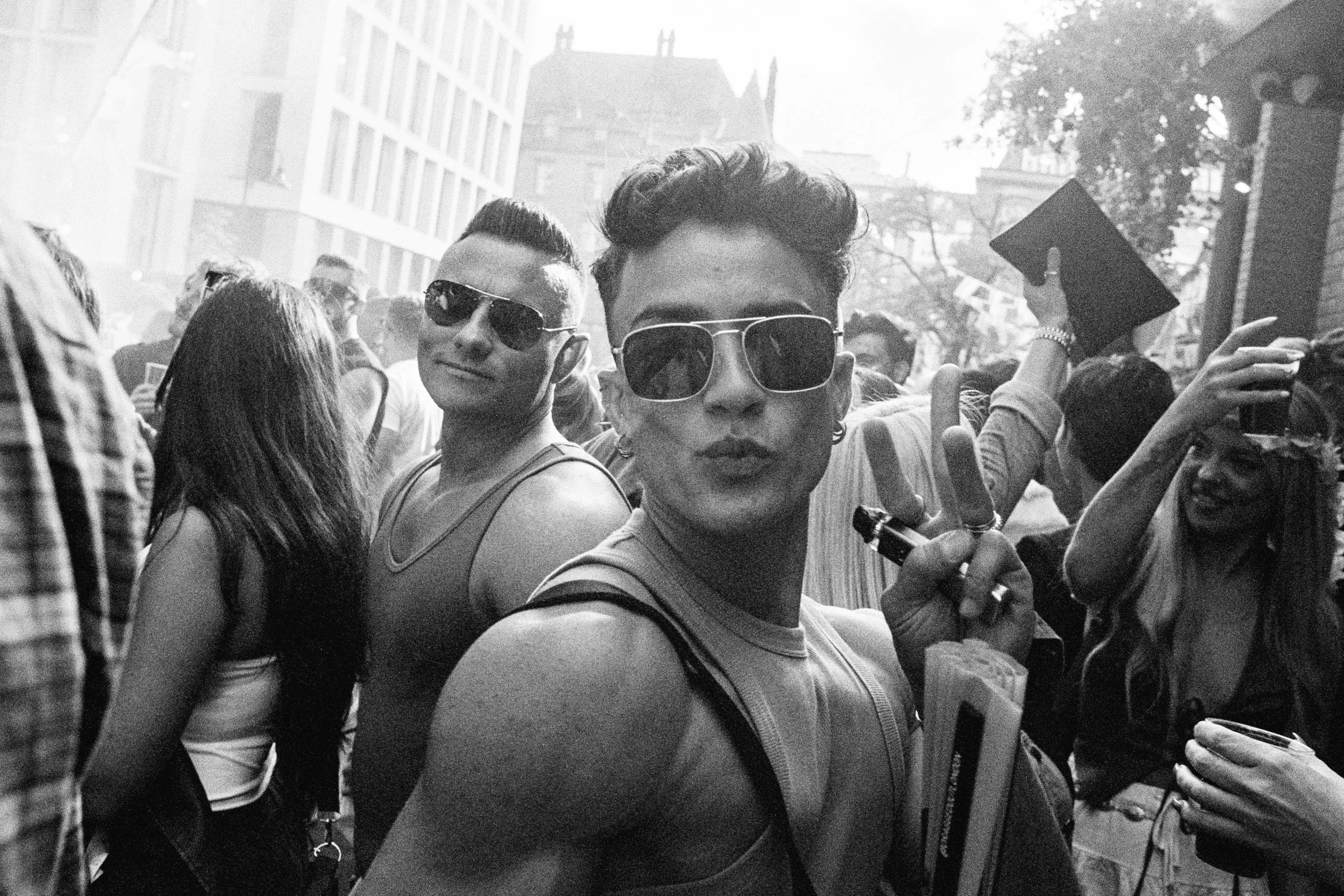 Black and white photo of two young men in sunglasses at a crowded outdoor event, one making a peace sign and the other smiling.