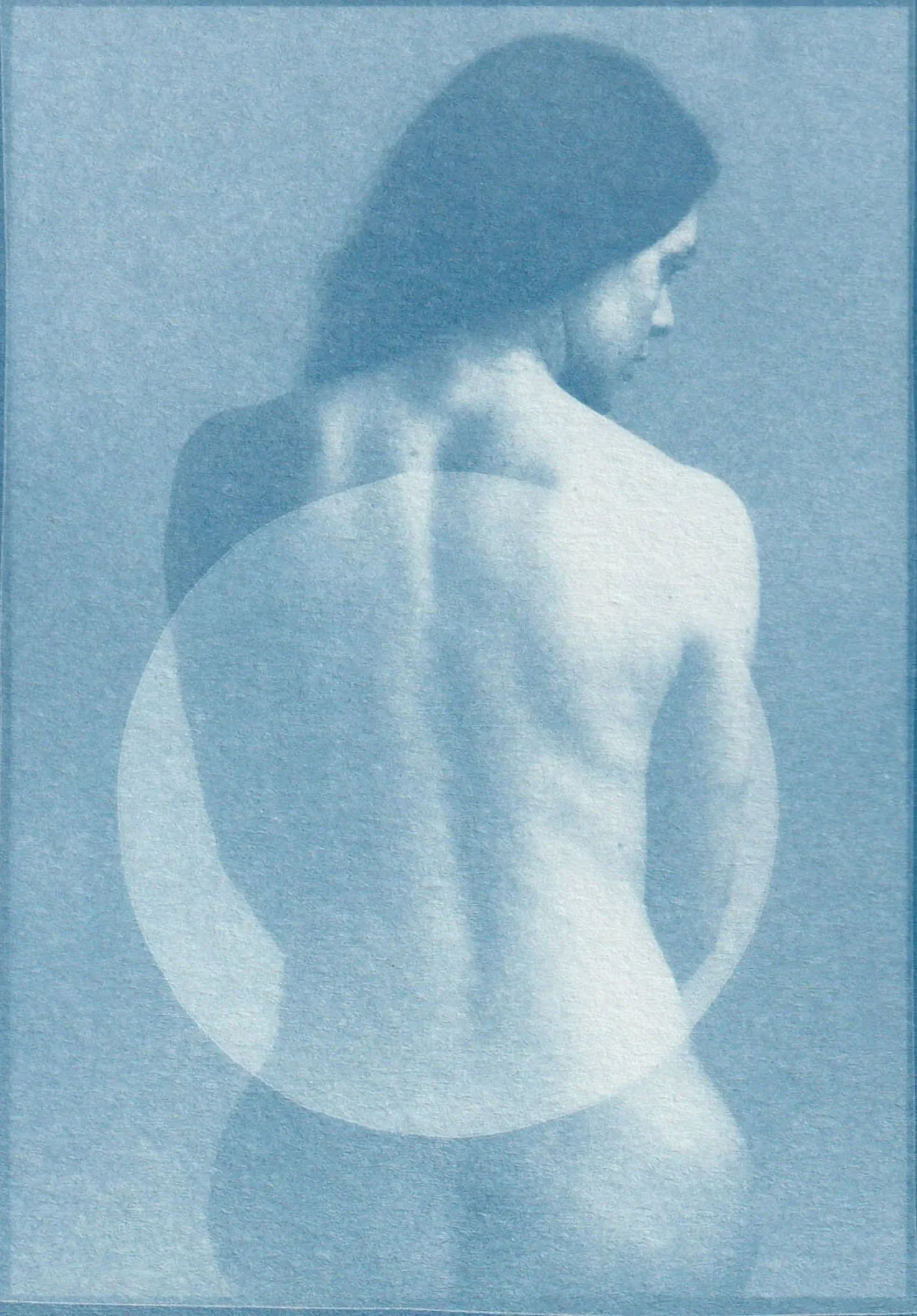 A black and white photograph of a topless woman with her back turned, looking down to her right, with shadows creating circles on the wall behind her.