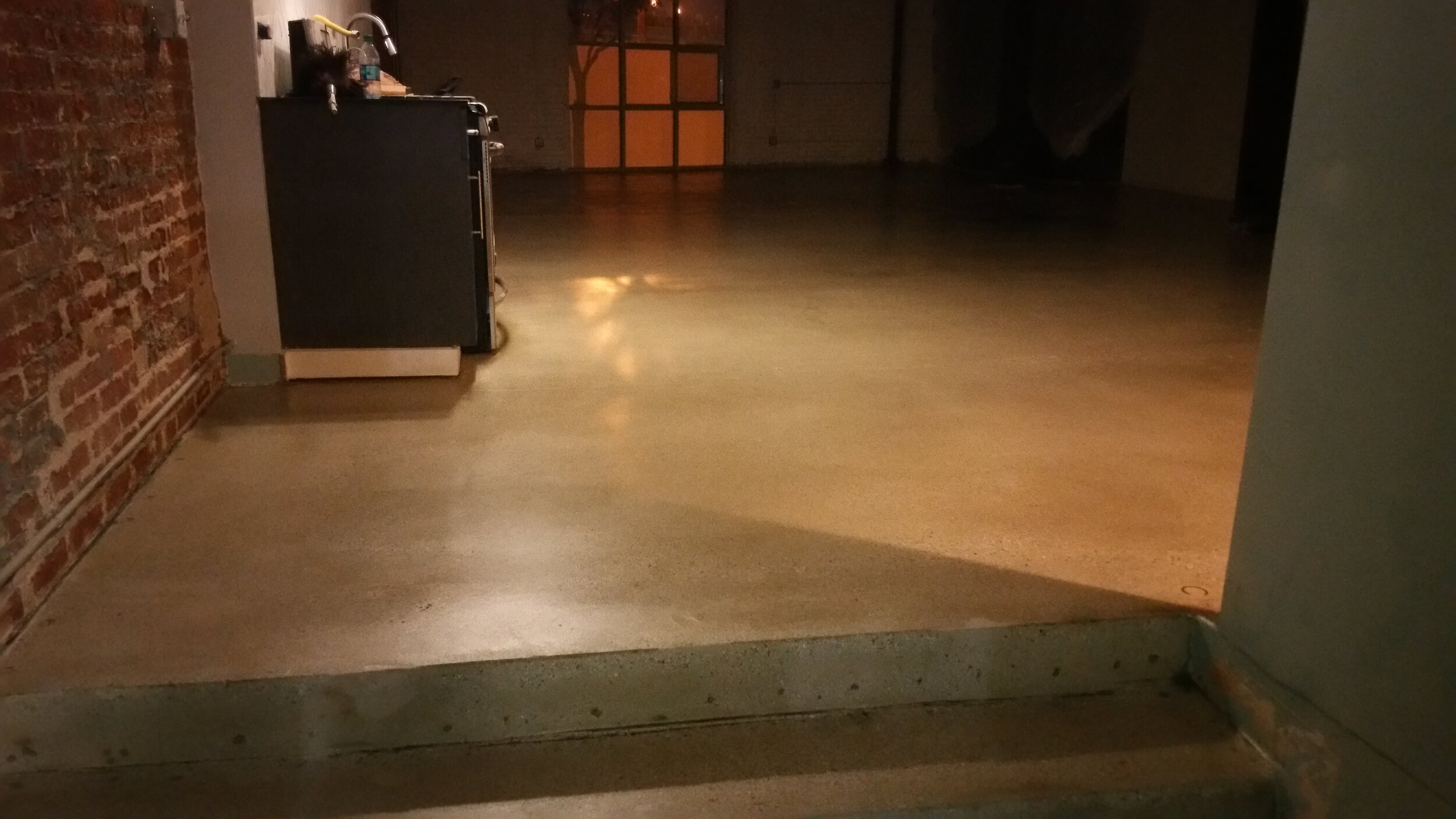 Empty indoor space with a brick wall on the left and a kitchen area, wood flooring, and a doorway in the background.