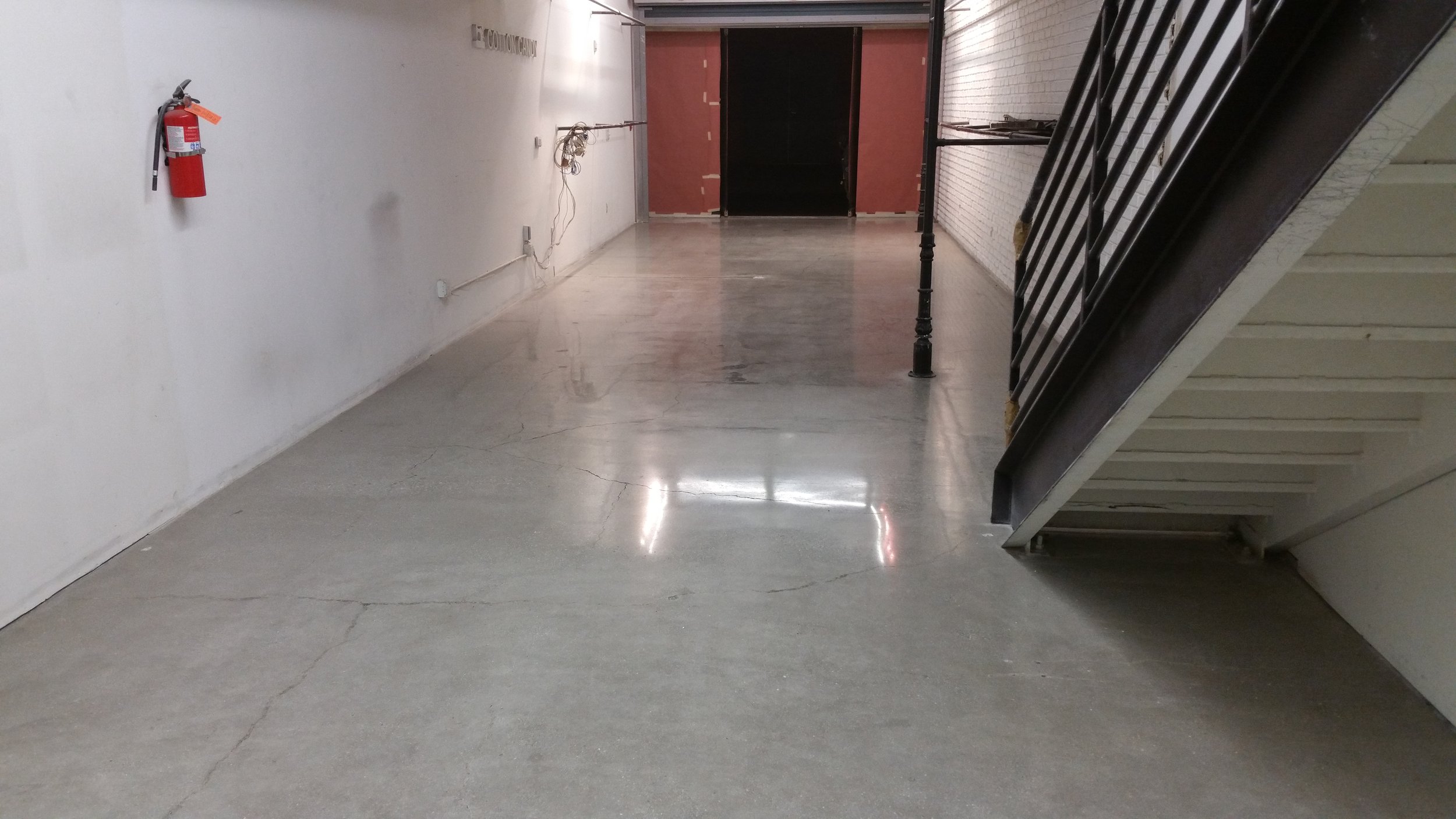 Empty hallway with concrete floor, white wall on the left with a fire extinguisher, visible wiring, and a staircase on the right side leading to an upper level.