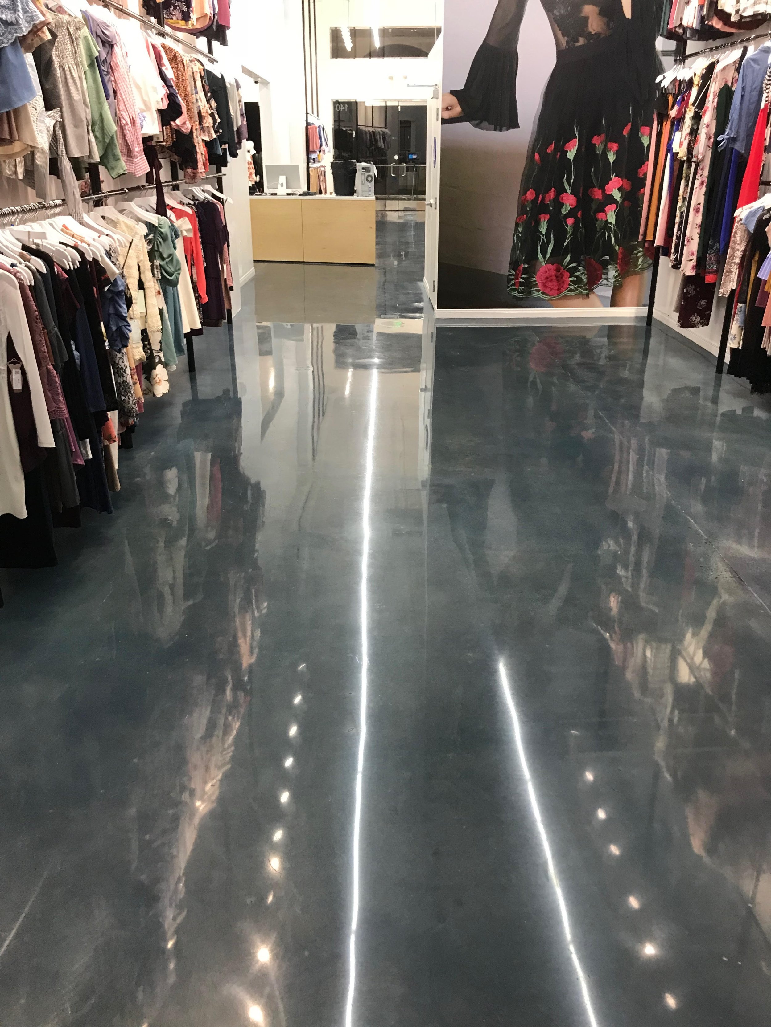 Empty clothing store aisle with racks of women's clothes on both sides and a large floral dress on a mannequin at the end of the aisle, with bright overhead lighting reflecting off the polished concrete floor.