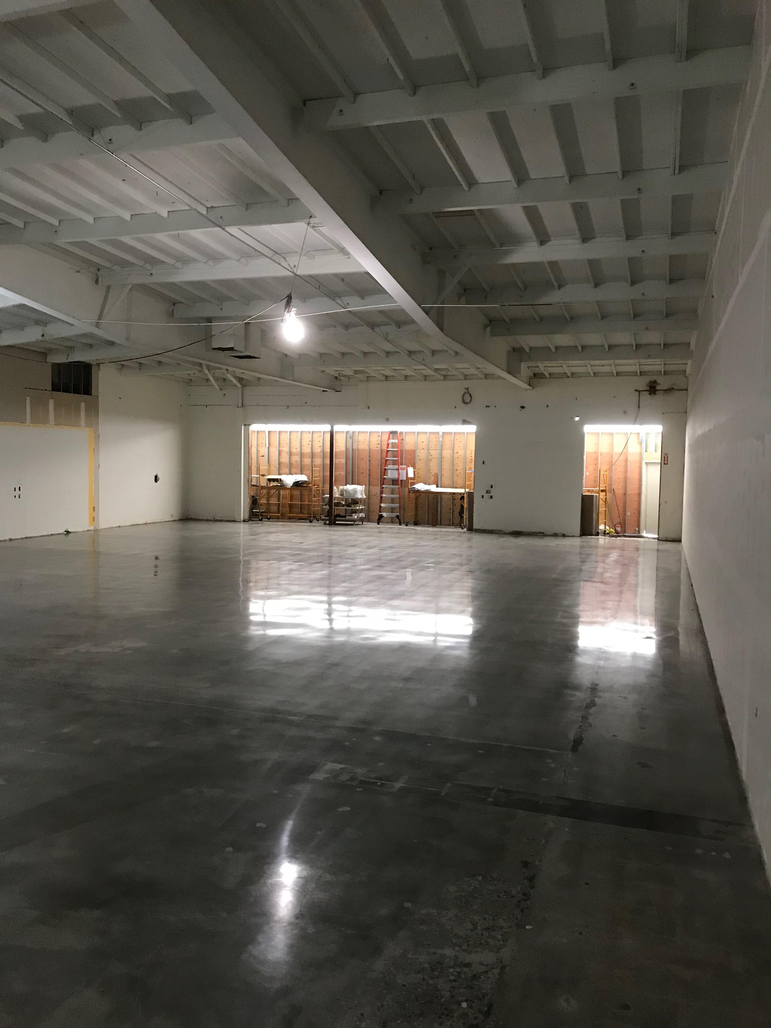 Empty indoor space under renovation with polished concrete floors, unfinished walls, exposed ceiling beams, and construction materials near open doorways.
