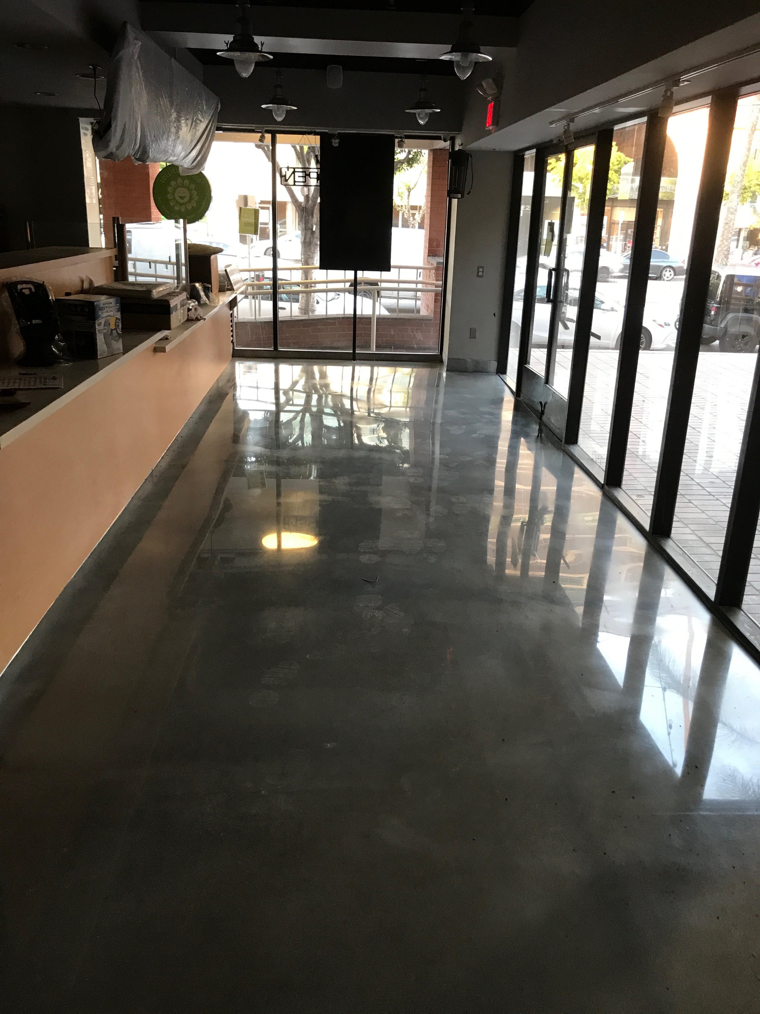 Empty restaurant with a counter on the left, large glass doors on the right, and sunlight coming through. The floor is polished concrete and reflects the light and surroundings.