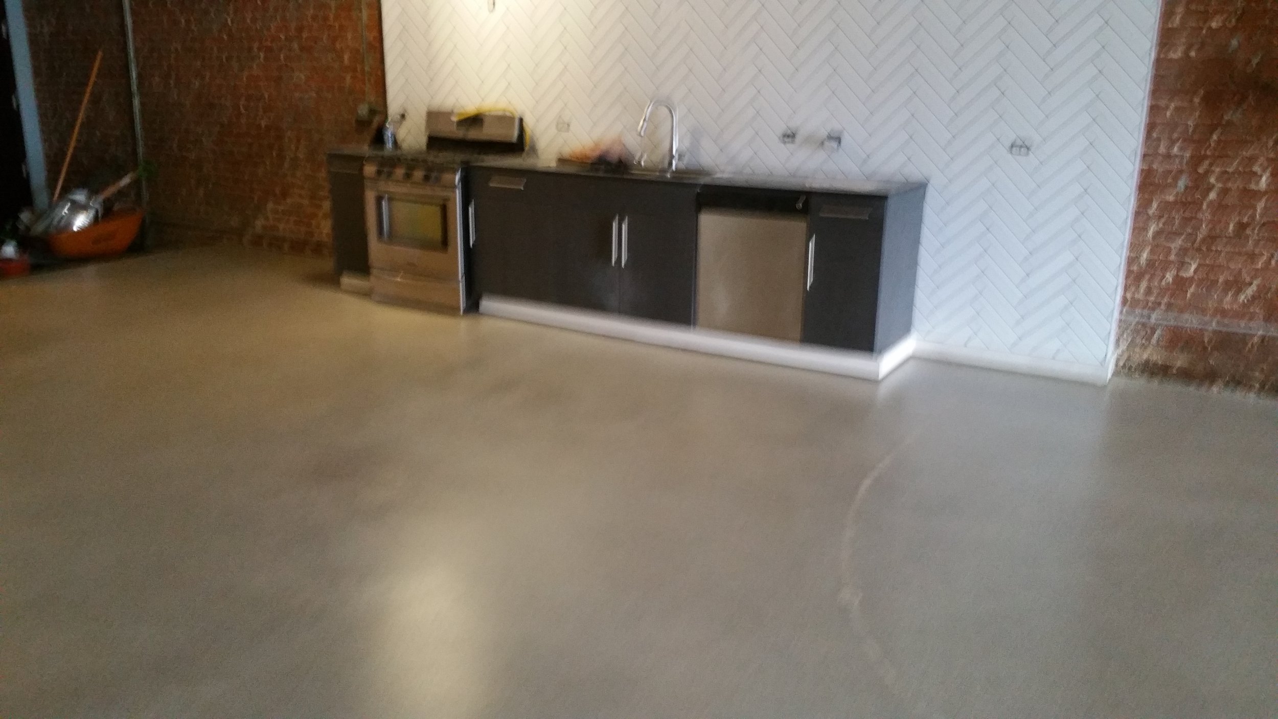 An industrial-style kitchen with a concrete floor, brick walls, and a black cabinetry along the wall with a sink and appliances.