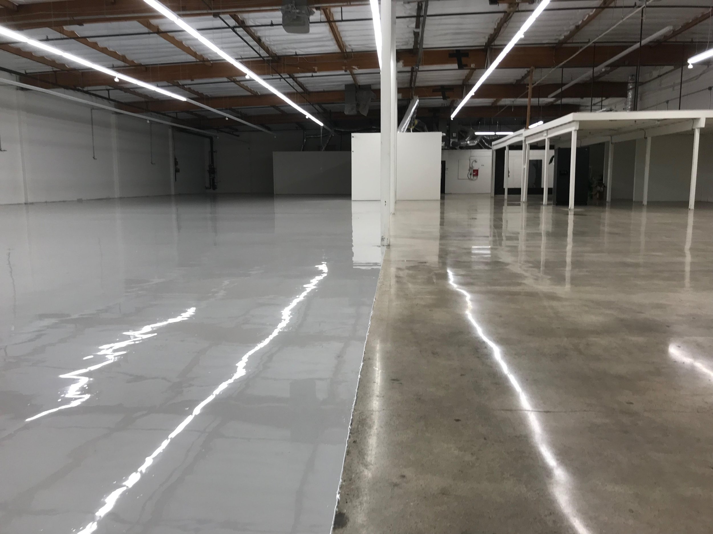 Industrial space with a sharp division between a glossy white floor on the left and a matte concrete floor on the right, separated by a vertical line. The room has exposed ceiling beams and multiple fluorescent light strips.